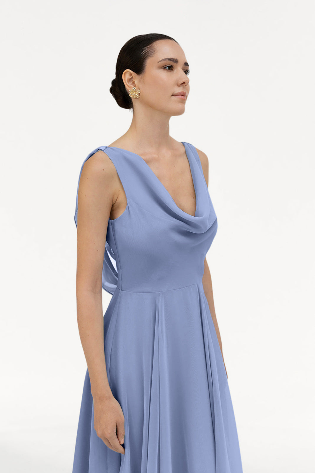 A woman with dark hair in a low bun wears the TH&TH Athena Bridesmaid Dress in French Blue Chiffon and gold earrings, standing against a plain white background.