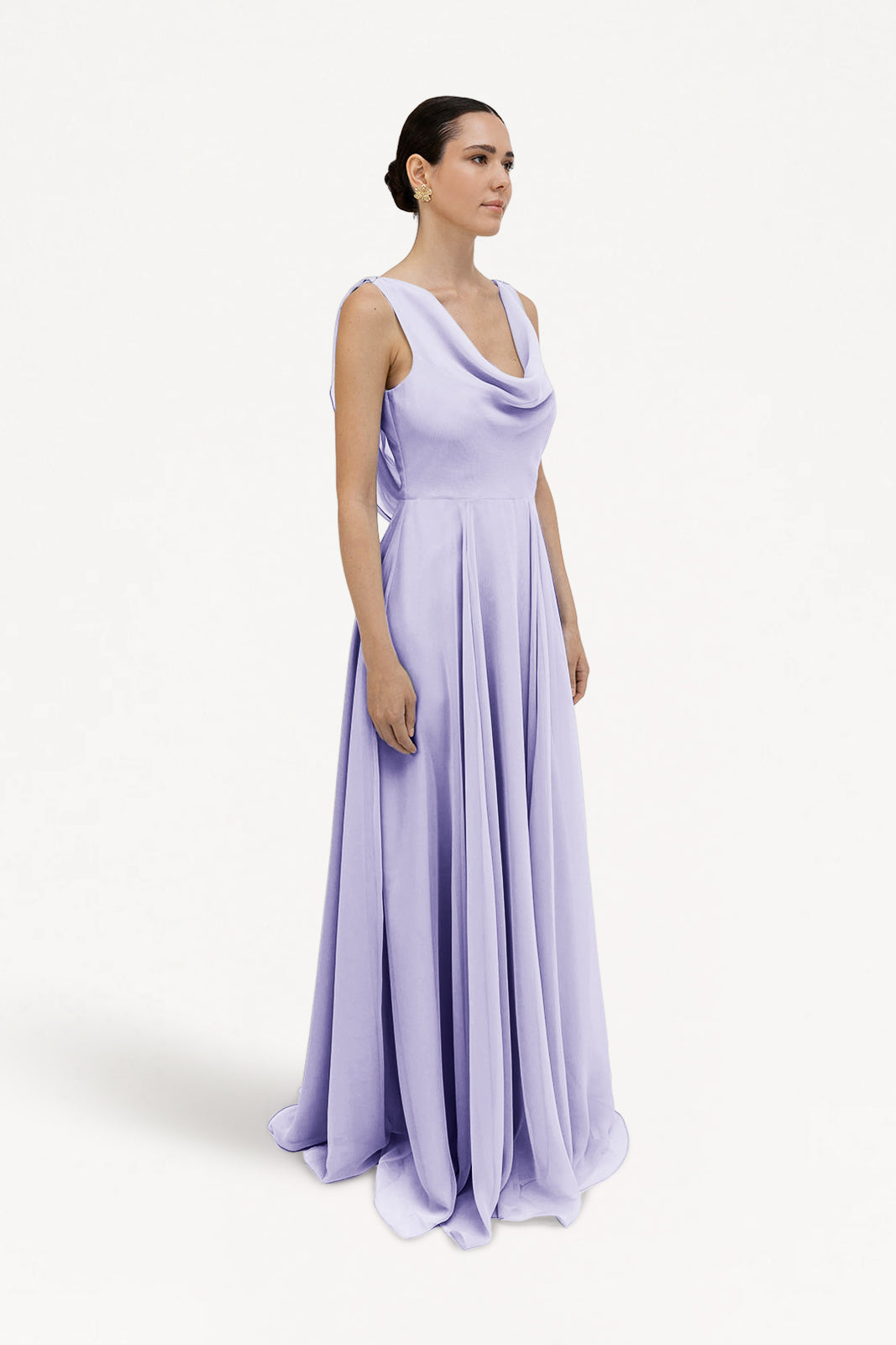 A woman in the TH&TH Athena Bridesmaid Dress in Lilac Mist Chiffon stands against a white background, her dark hair pulled back in a sleek bun as she looks to the side. The dress features a long, flowing silhouette and draped neckline.