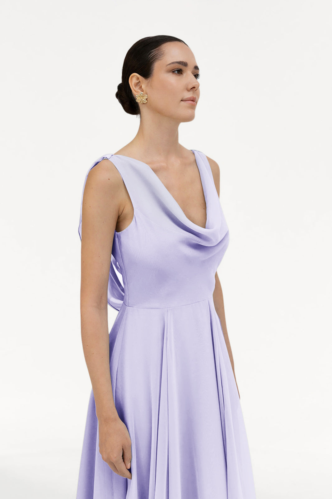A woman with dark hair in a low bun wears the TH&TH Athena Bridesmaid Dress in Lilac Mist chiffon and gold earrings, standing against a plain white background.