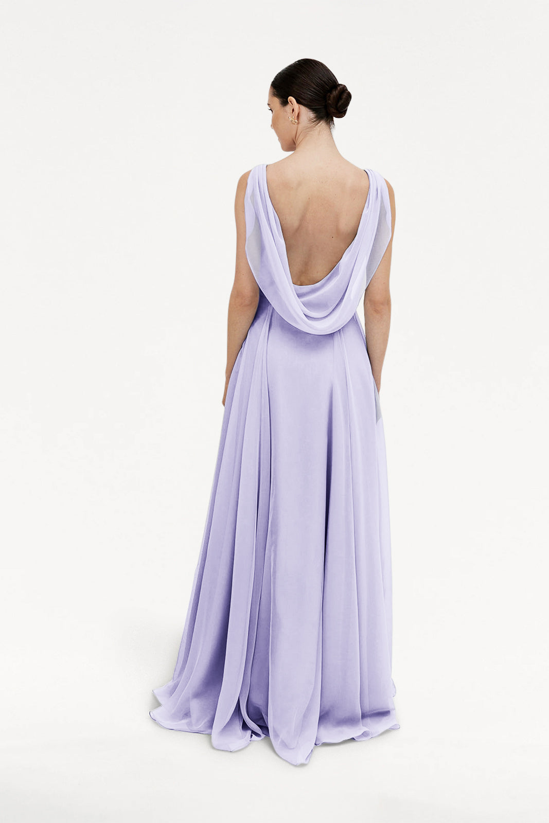A woman with dark hair in a bun stands facing away, wearing the TH&TH Athena Bridesmaid Dress in Lilac Mist chiffon, featuring a flowing skirt and draped low cowl back, set against a plain white background.