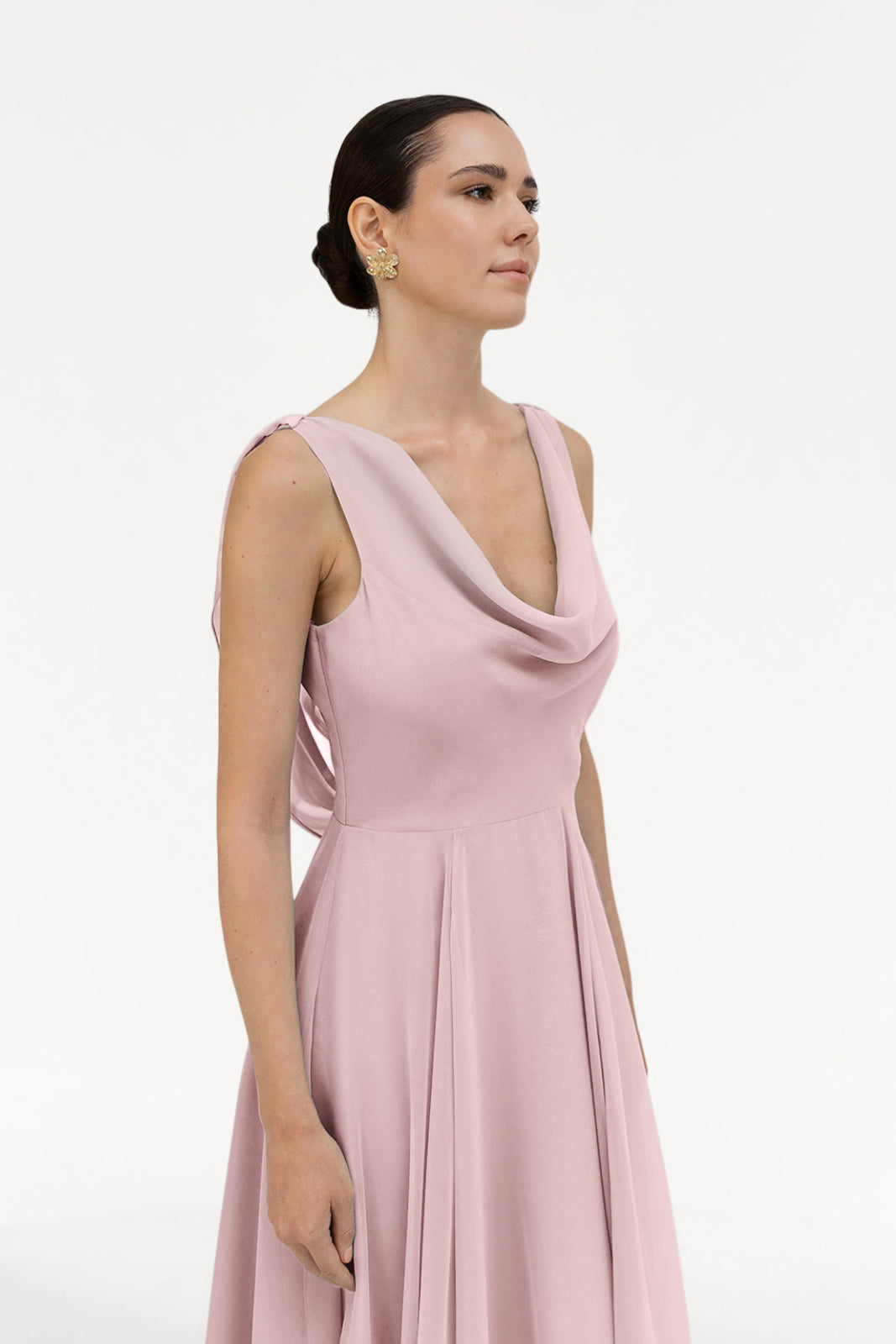 A woman with dark hair in a low bun wears the TH&TH Athena Bridesmaid Dress in smoked blush chiffon and gold earrings, standing against a plain white background and looking slightly to the side.