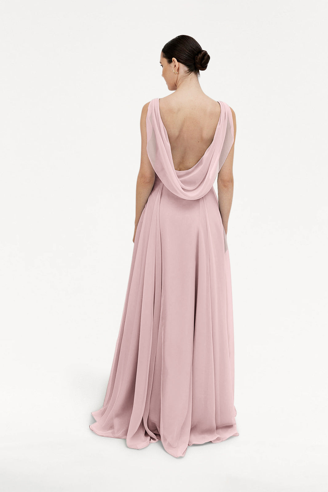 A woman with dark hair in a bun stands facing away against a white background, wearing the TH&TH Athena Bridesmaid Dress in Smoked Blush Chiffon, featuring a long, flowing skirt and draped low-cut back.