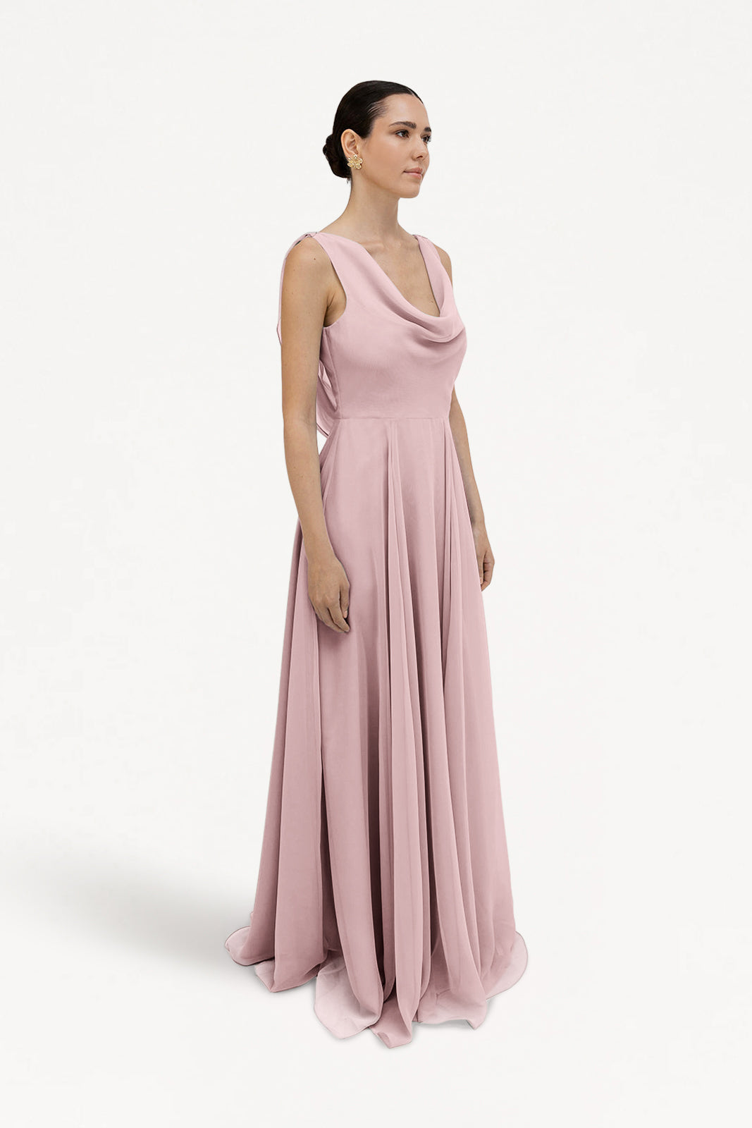 A woman wears the Athena Bridesmaid Dress in Smoked Blush Chiffon by TH&TH, featuring a sleeveless, floor-length design with a draped cowl neck and flowing skirt. She stands against a plain white background, hair pulled back, facing slightly right.