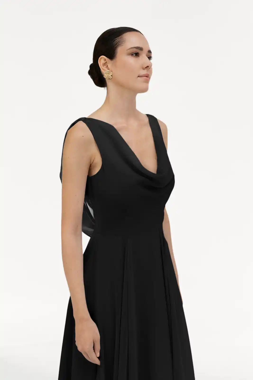 A woman with dark hair in a low bun wears the Athena Bridesmaid Dress in Black Chiffon by TH&TH and gold earrings, standing against a plain white background.