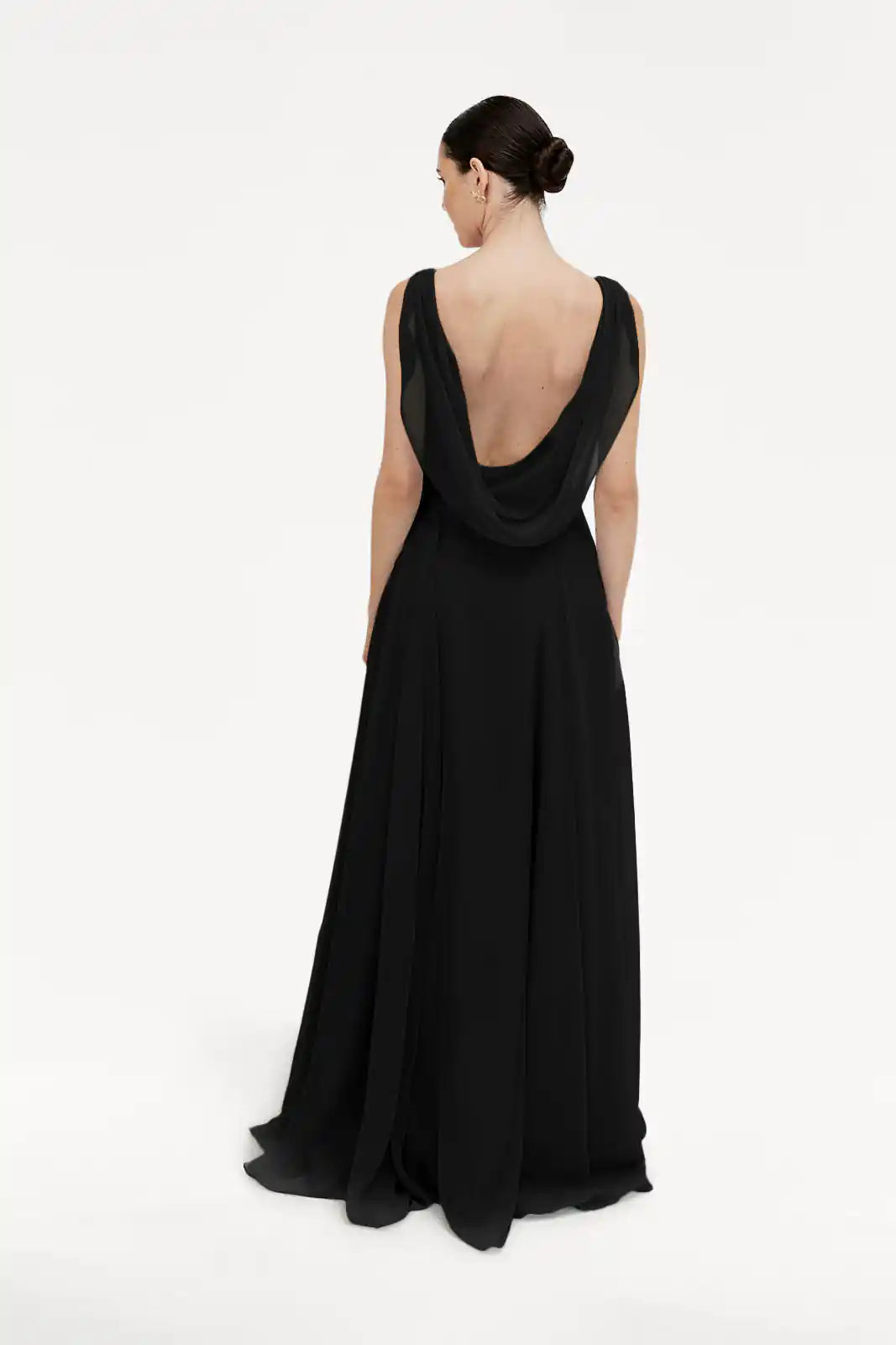 A woman with her hair in a neat bun faces away, wearing the TH&TH Athena Bridesmaid Dress in black chiffon—a long, flowing gown with a cowl neck and deep draped open back—set against a plain white background.