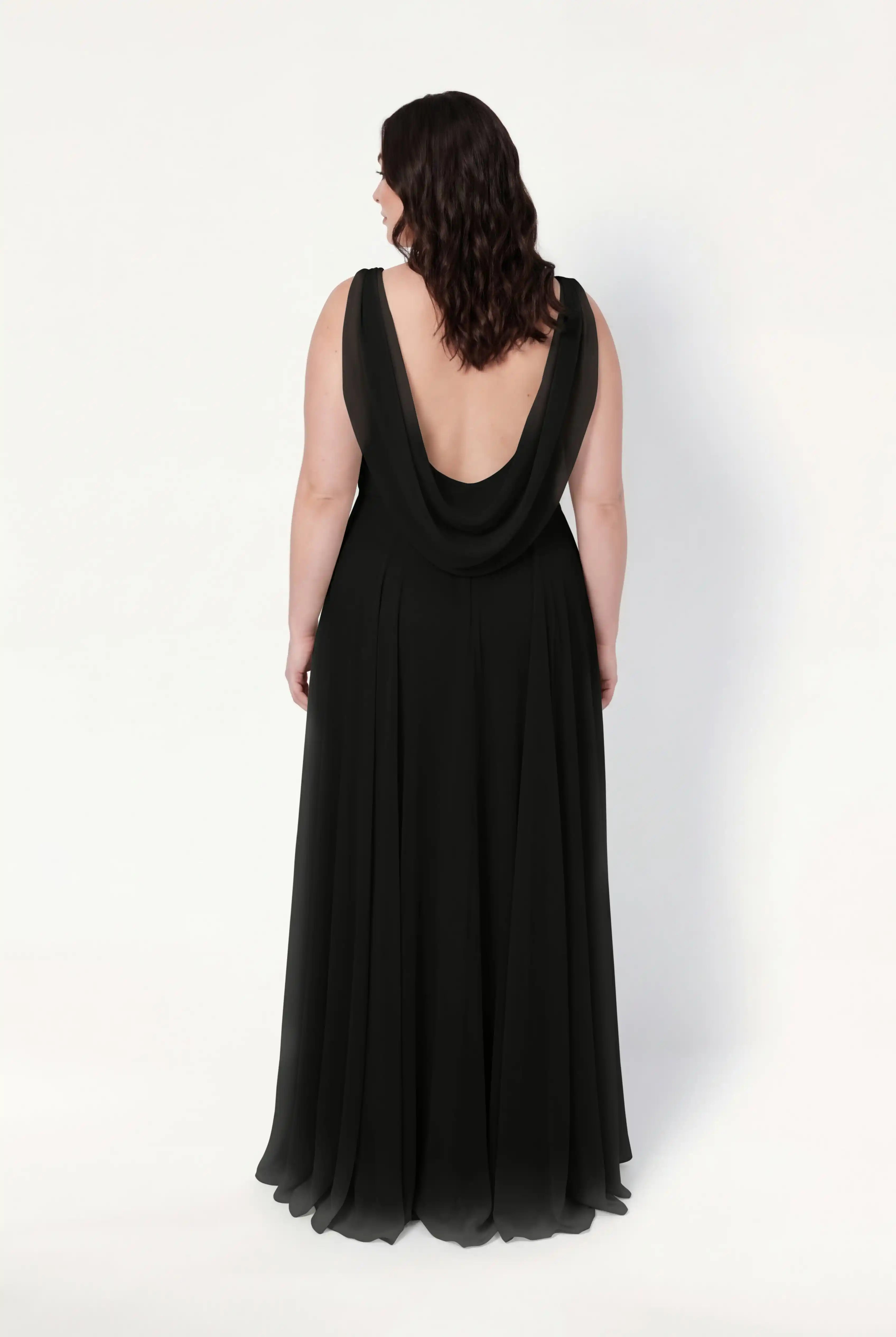 A woman with wavy dark hair is seen from behind wearing the TH&TH Athena Bridesmaid Dress in black chiffon, featuring a long, flowing silhouette and deep draped back, set against a plain white background.