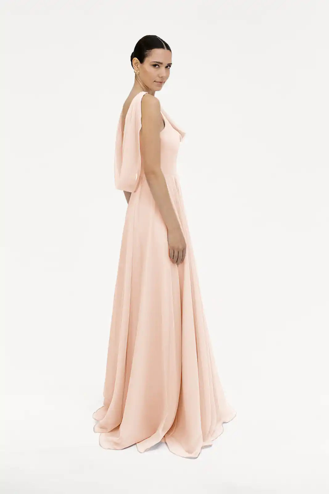 A woman wears the TH&TH Athena Bridesmaid Dress in blush pink chiffon, a floor-length gown with draped sleeves and a cowl neck, posing against a plain white background.