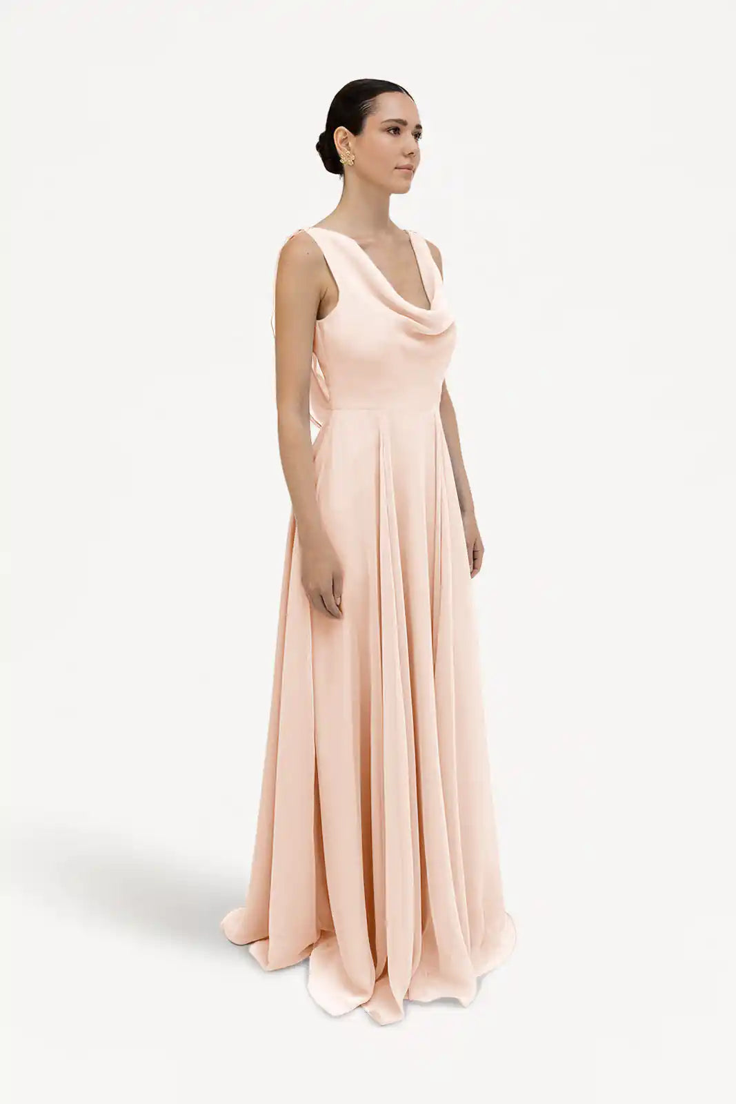 A woman with slicked-back hair stands against a white background, wearing the TH&TH Athena Bridesmaid Dress in blush pink chiffon—a sleeveless, floor-length gown featuring a draped cowl neck.