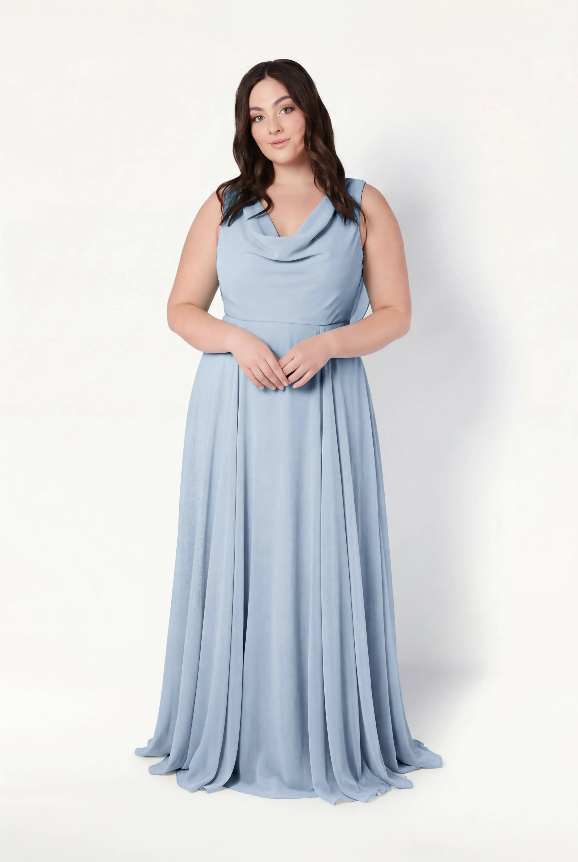 A woman with long, wavy dark hair smiles gently at the camera, wearing the TH&TH Athena Bridesmaid Dress in Dusty Blue Chiffon—a floor-length, sleeveless gown with a draped cowl neck and flowing skirt against a light background.