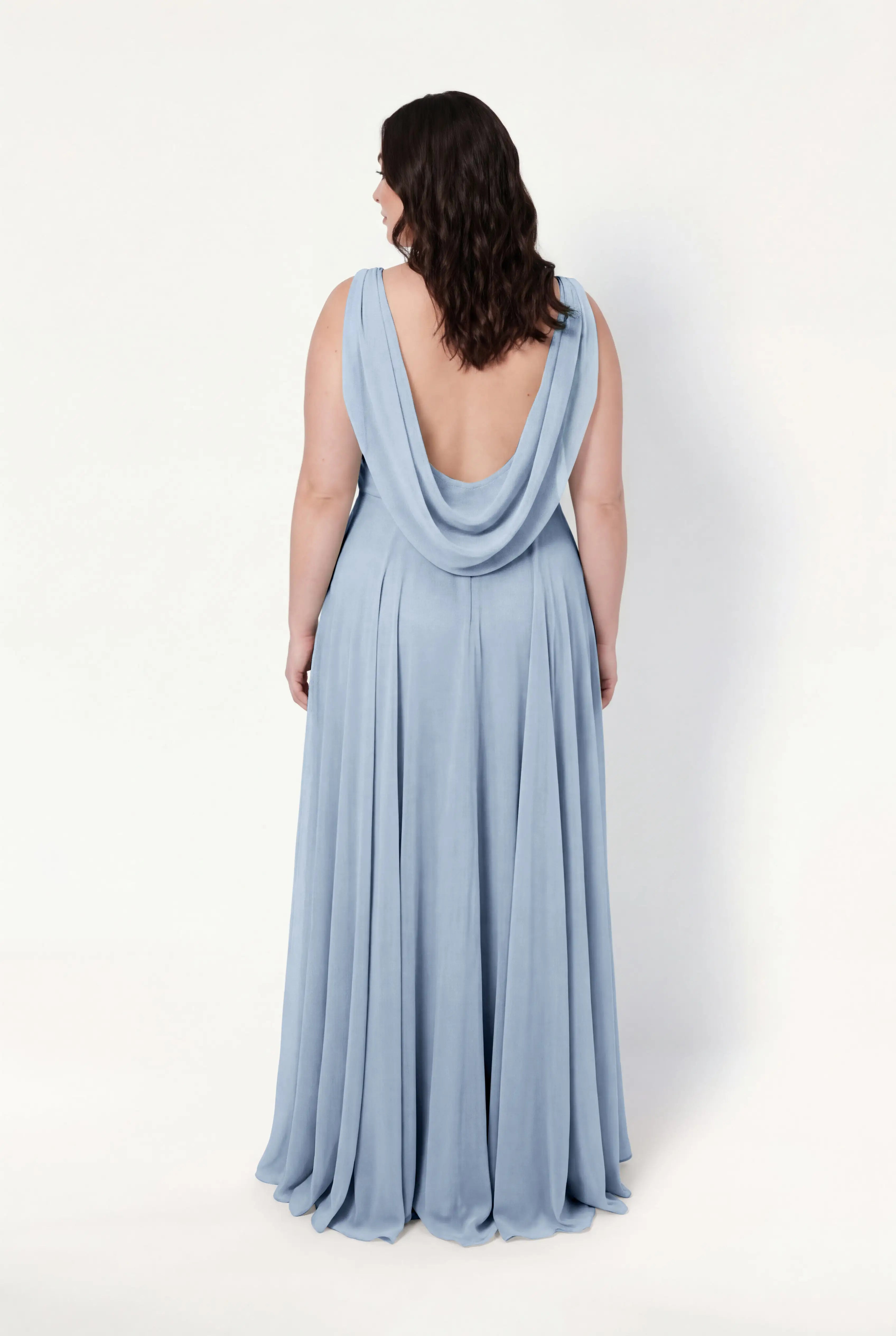 A woman with wavy dark hair models the TH&TH Athena Bridesmaid Dress in Dusty Blue Chiffon, showing off its flowing fabric and draped low open-back design as she stands facing away against a plain white background.