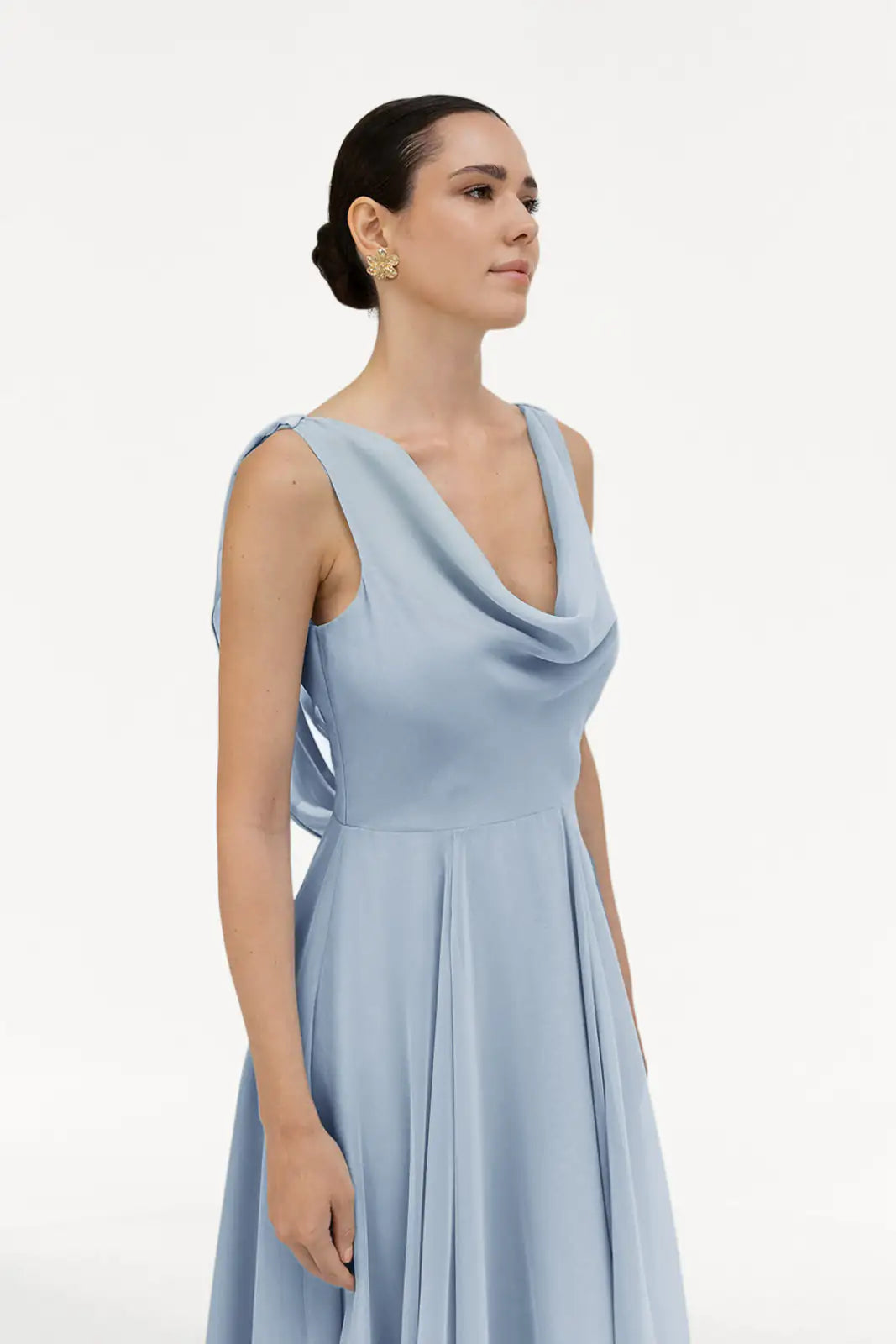 A woman with dark hair in a low bun wears the TH&TH Athena Bridesmaid Dress in Dusty Blue Chiffon and gold earrings, standing against a plain white background.