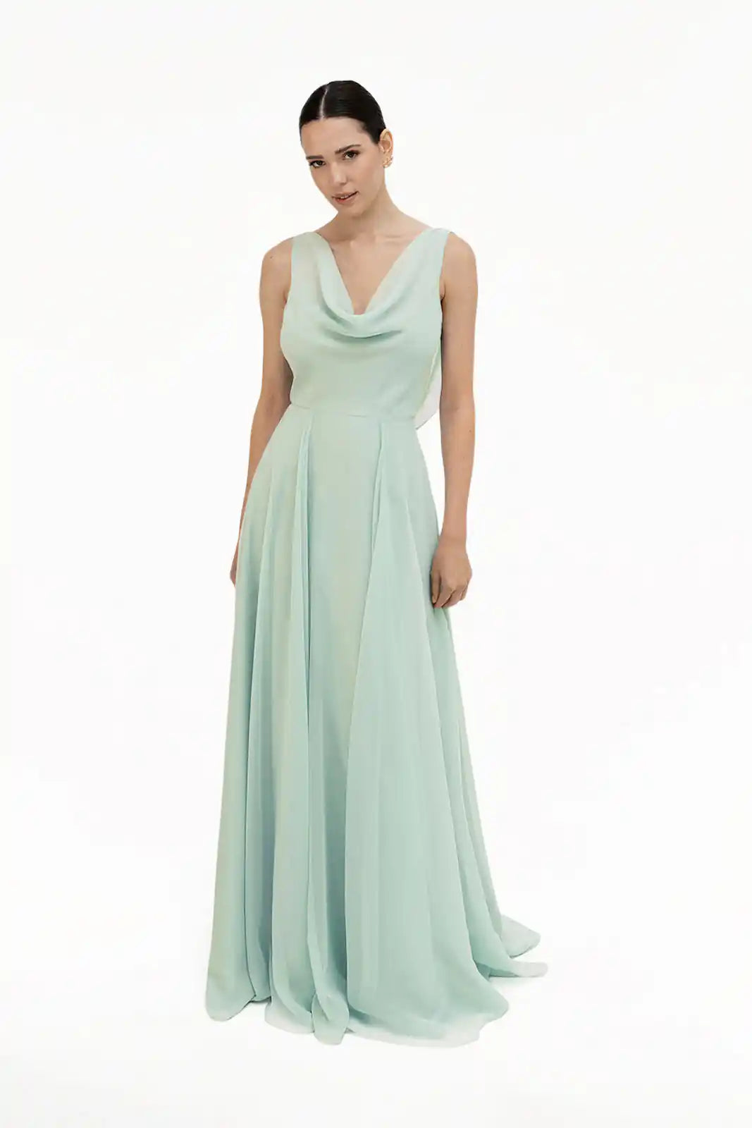 A woman poses against a plain white background in the Athena Bridesmaid Dress in Dusty Eucalyptus by TH&TH—a sleeveless, floor-length chiffon gown with a draped cowl neck. Her dark hair is pulled back.