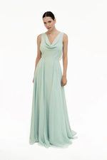 A woman poses against a plain white background in the Athena Bridesmaid Dress in Dusty Eucalyptus by TH&TH—a sleeveless, floor-length chiffon gown with a draped cowl neck. Her dark hair is pulled back.