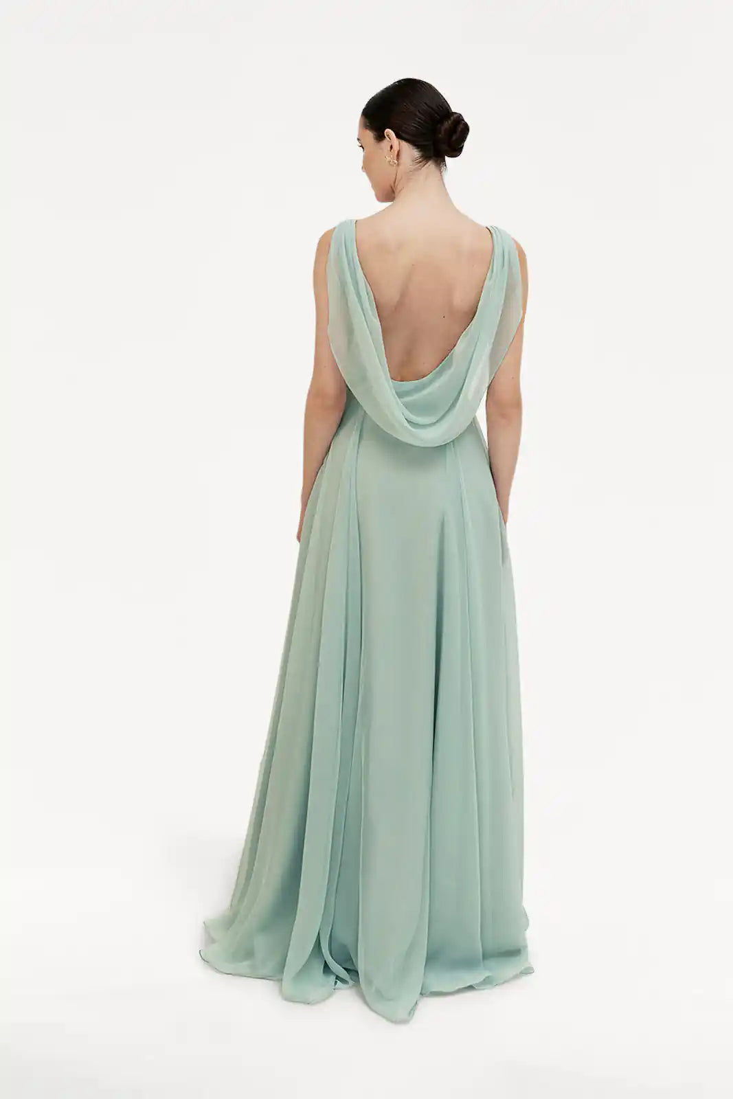 A woman faces away, wearing the TH&TH Athena Bridesmaid Dress in Dusty Eucalyptus—a flowing, floor-length chiffon gown with a draped cowl neck and low back. Her hair is neatly styled in a bun, set against a plain white background.