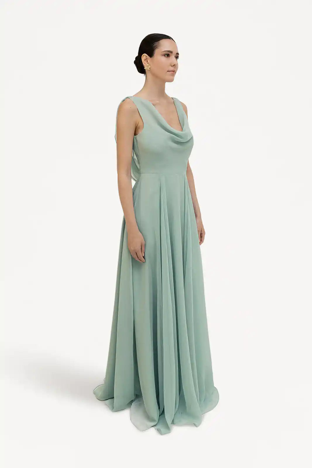 A woman with dark hair pulled back wears the TH&TH Athena Bridesmaid Dress in Dusty Eucalyptus, featuring a long, flowing chiffon silhouette and draped cowl neck, standing against a plain white background looking slightly to the side.