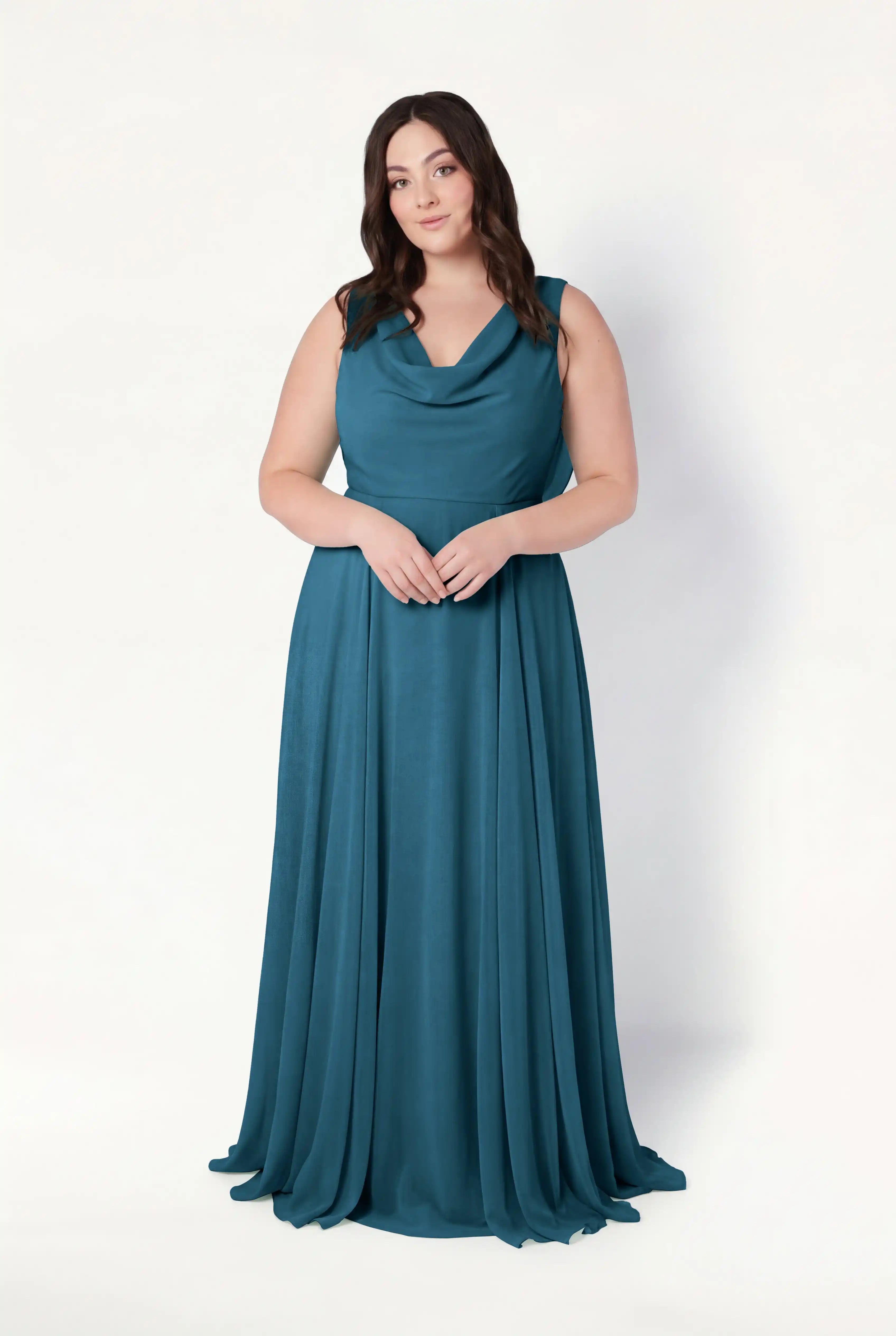 A woman with long brown hair poses against a white background in the TH&TH Athena Bridesmaid Dress in Emerald Dawn, featuring a draped neckline and flowing skirt. Her hands are clasped, expression neutral, exuding timeless elegance.