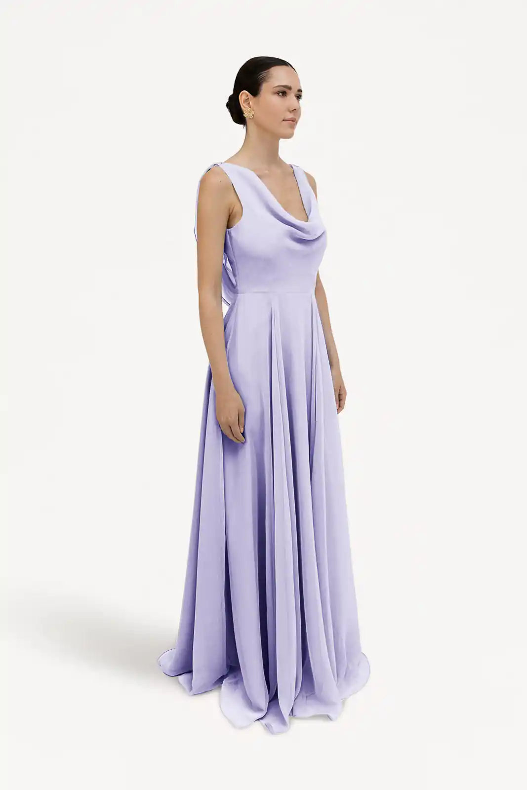 A woman in the TH&TH Athena Bridesmaid Dress in Lilac Mist Chiffon stands against a white background, her dark hair pulled back in a sleek bun as she looks to the side. The dress features a long, flowing silhouette and draped neckline.