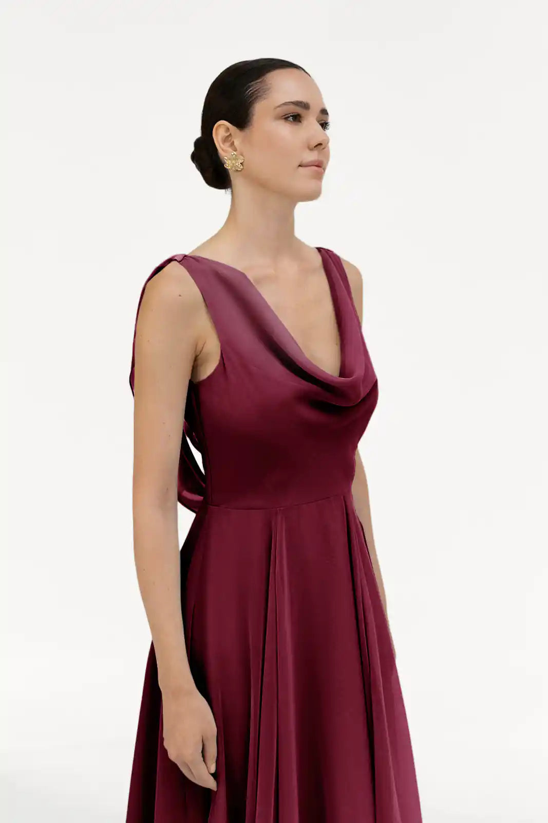 A woman wearing the TH&TH Athena Bridesmaid Dress in Merlot chiffon, a sleeveless gown with a cowl neck and open back, poses against a white background. Her hair is styled in a neat bun and she wears gold earrings.