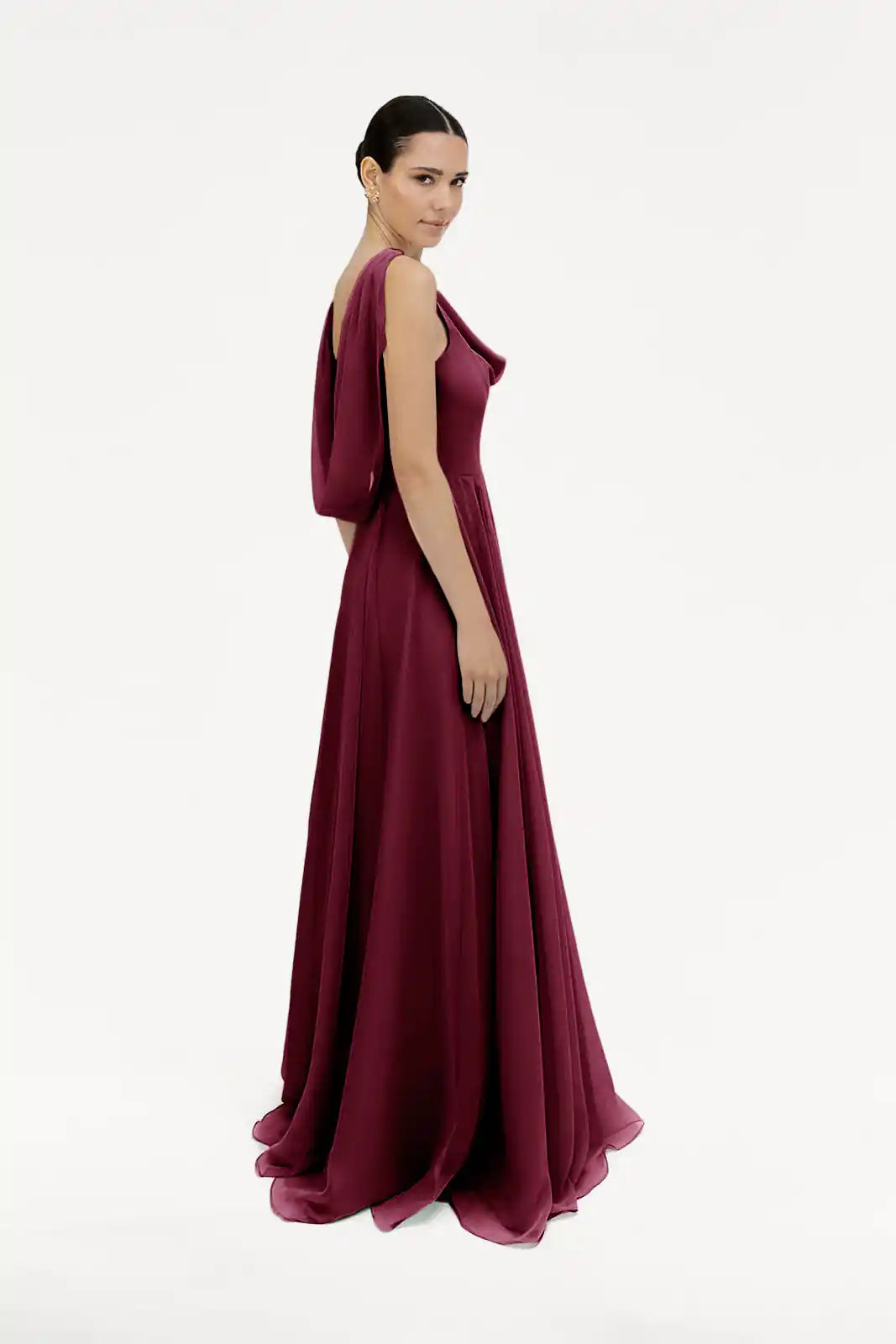 A woman stands sideways against a white background, wearing the TH&TH Athena Bridesmaid Dress in Merlot Chiffon—a flowing, sleeveless, floor-length gown—looking towards the camera with a calm expression.