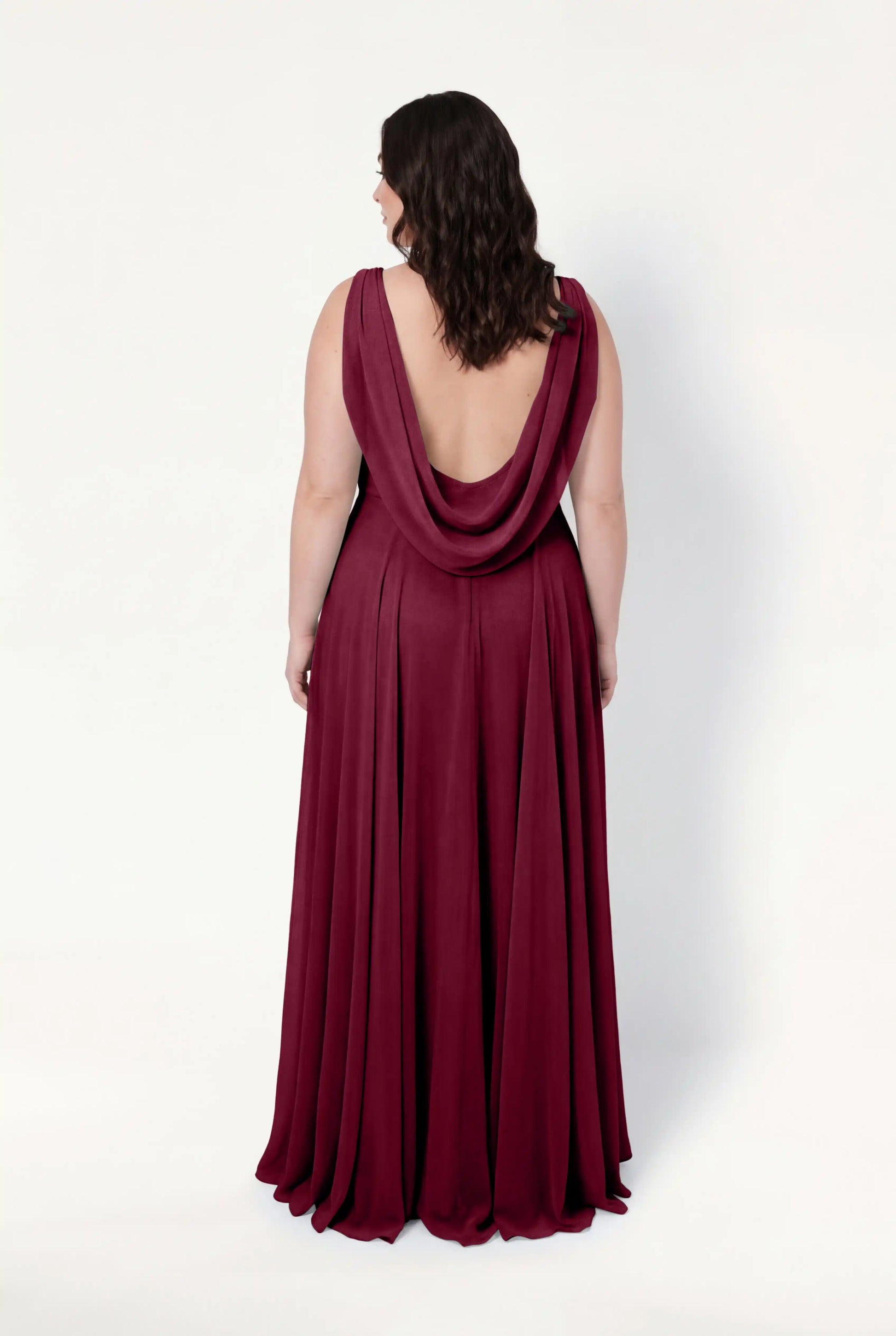 A woman with wavy brown hair faces away, wearing the TH&TH Athena Bridesmaid Dress in Merlot Chiffon—a sleeveless, floor-length gown with a draped open back—set against a plain white background.