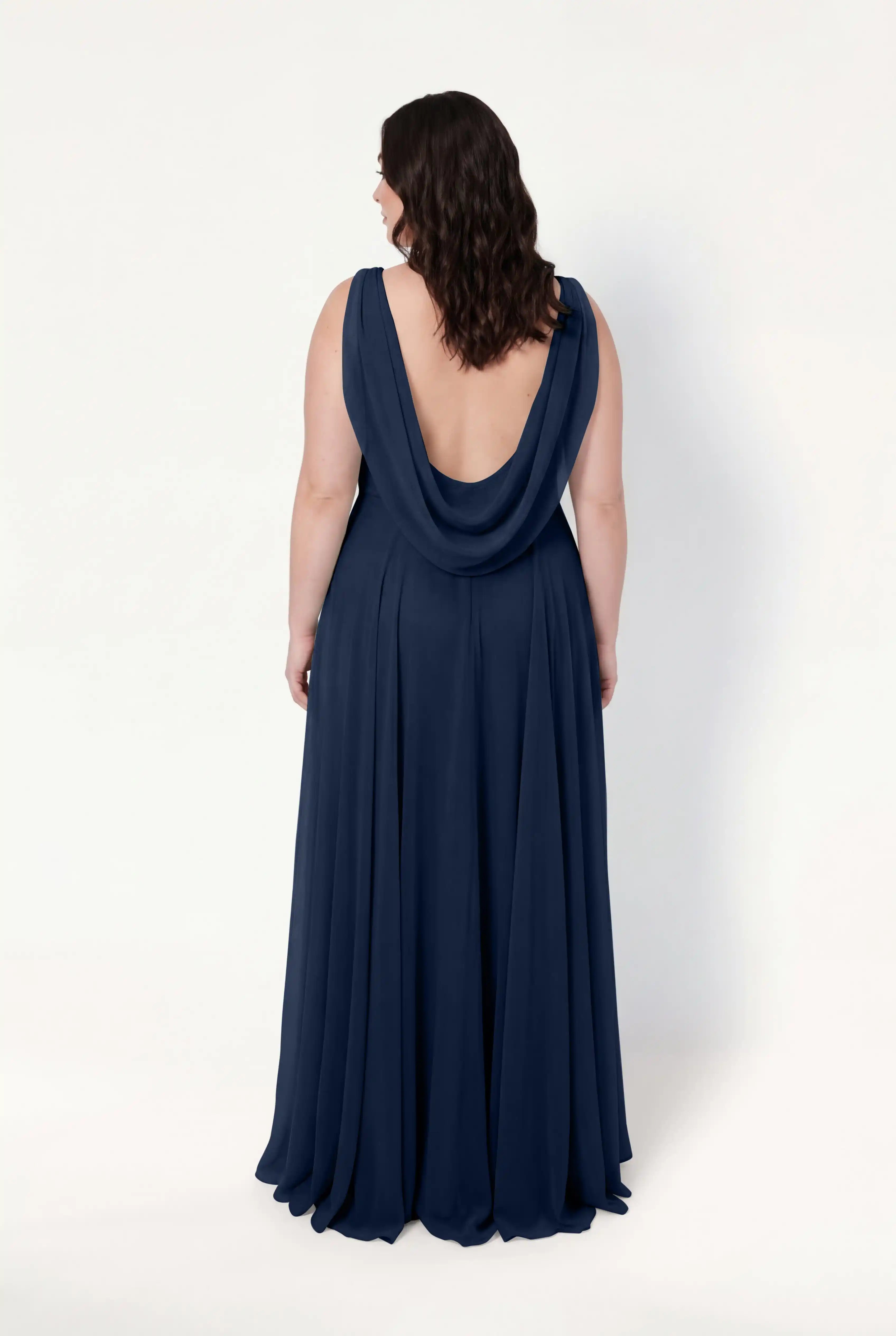 A woman with wavy brown hair models the TH&TH Athena Bridesmaid Dress in navy chiffon, featuring a draped cowl neck and low-cut back, standing with her back to the camera against a plain white background.