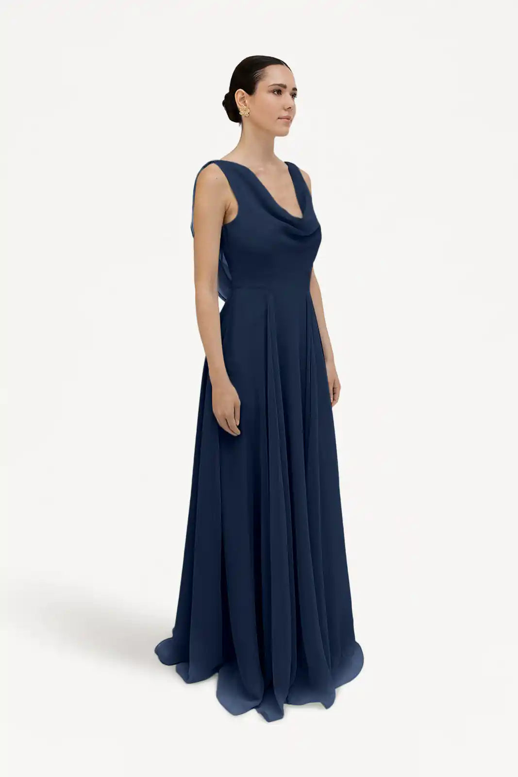 A woman poses against a white background in the TH&TH Athena Bridesmaid Dress in navy chiffon, featuring a sleeveless, floor-length design with a cowl neck. Her hair is pulled back as she gazes slightly to the side.