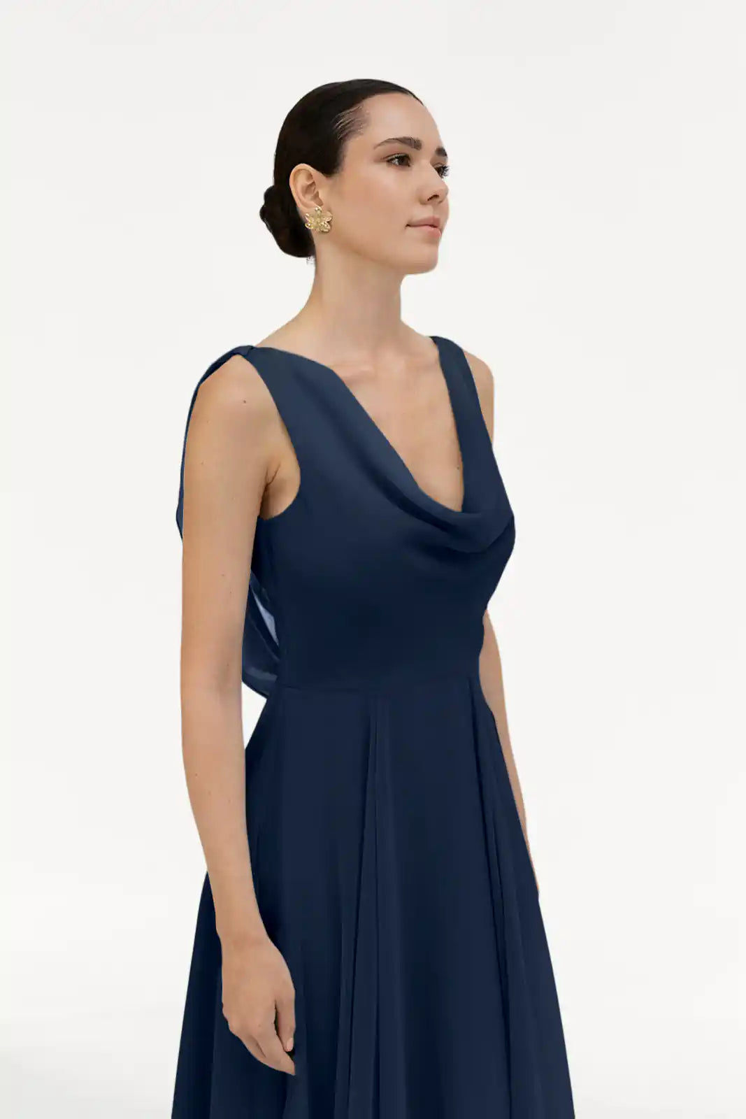 A woman with dark hair in a low bun models the TH&TH Athena Bridesmaid Dress in navy chiffon, featuring a sleeveless design and draped cowl neck, standing against a plain white background with a soft, sideways glance.