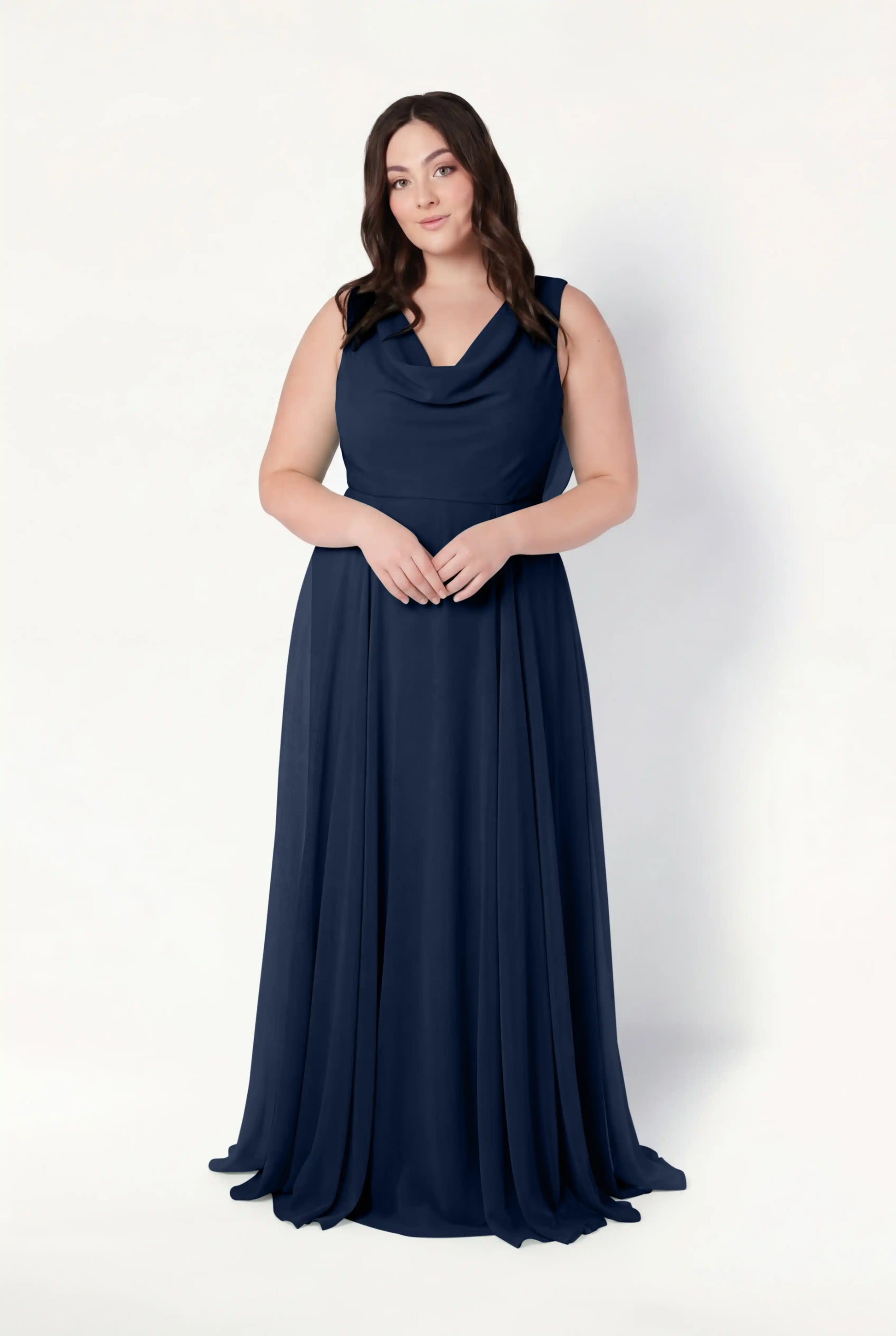 A woman with long brown hair stands against a white background, wearing the TH&TH Athena Bridesmaid Dress in navy chiffon. The sleeveless, floor-length gown features a draped cowl neck as she faces forward with her hands clasped gently together.