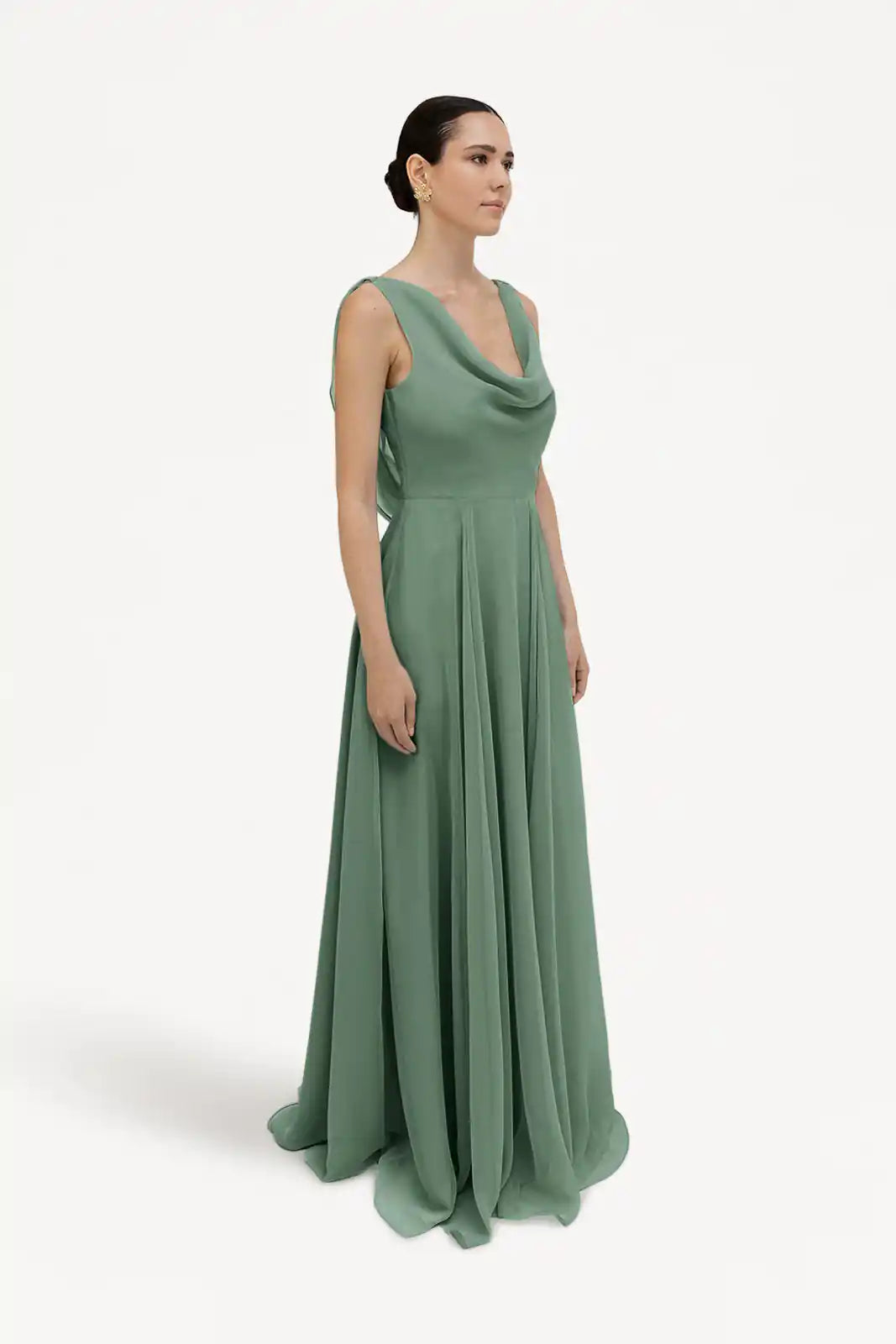 A woman, hair in a low bun, models the TH&TH Athena Bridesmaid Dress in sage green chiffon—a sleeveless, floor-length gown with a draped cowl neck—standing slightly turned against a plain white background.