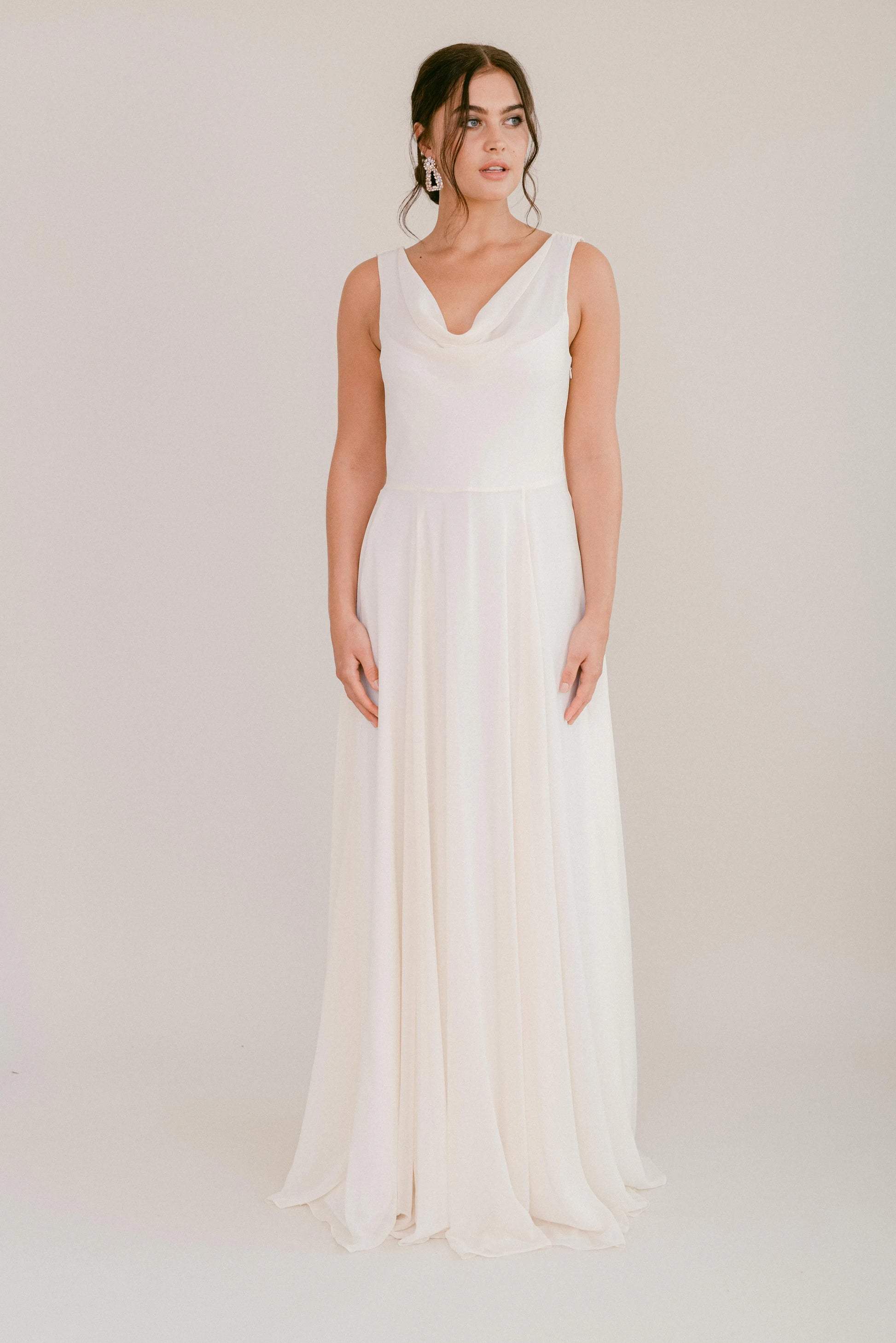 A woman poses against a plain background in the Athena Dress in Ivory Chiffon by TH&TH—a sleeveless, floor-length gown with a draped neckline and flowing skirt. Her hair is styled in a loose updo and she wears a calm expression.