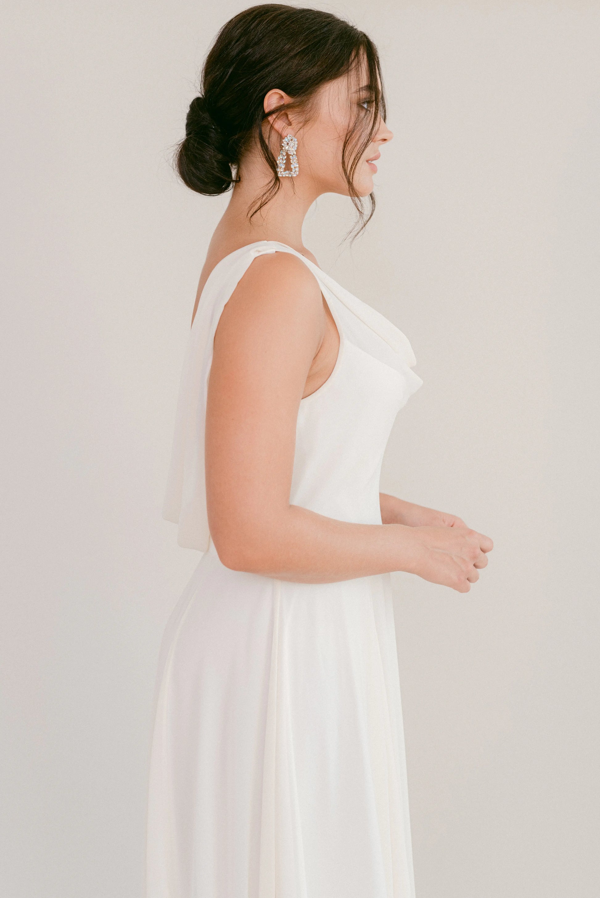 A woman with dark hair in a low bun models the TH&TH Athena Dress in Ivory Chiffon, shown in profile against a plain background and accessorized with statement drop earrings.