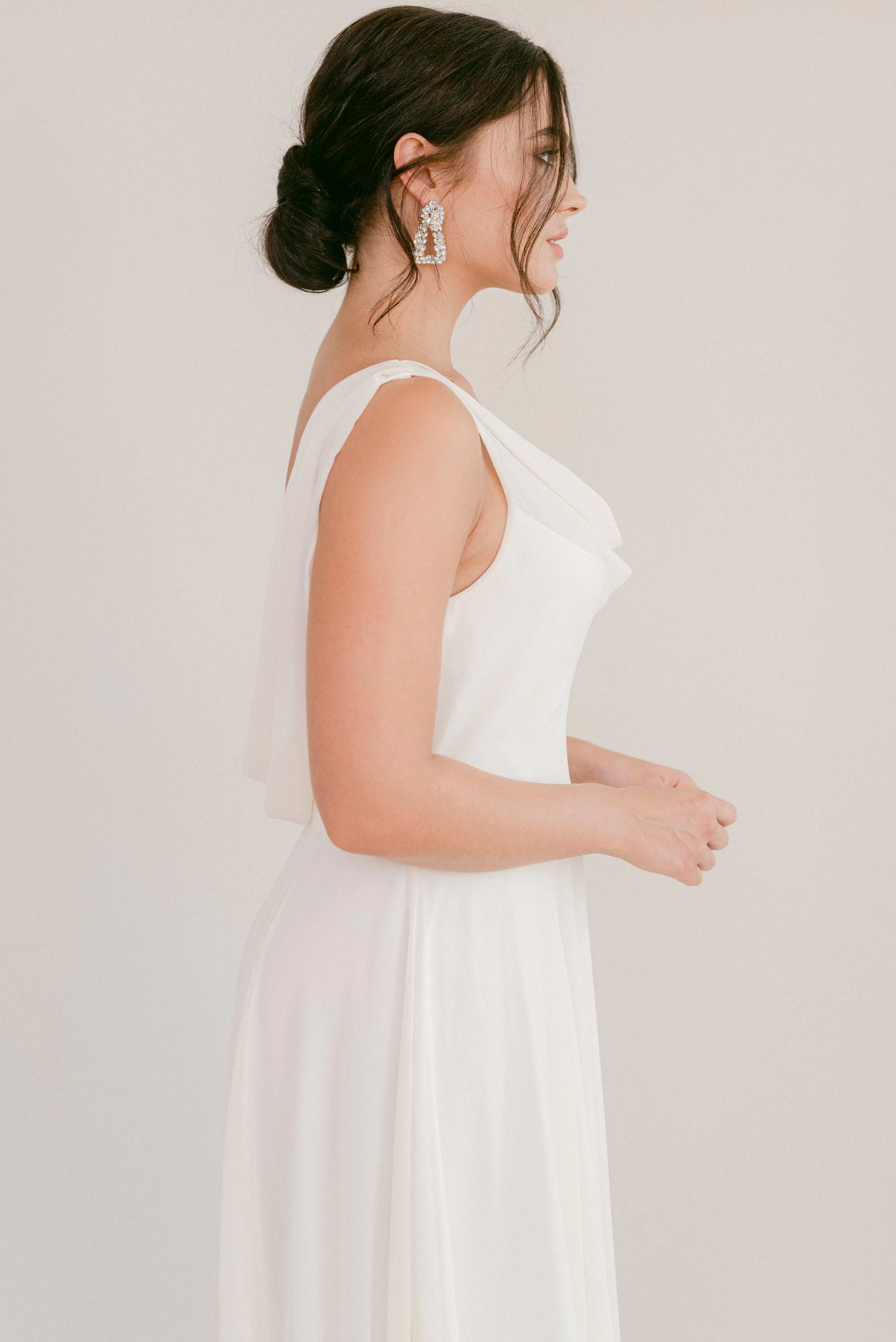 A woman with dark hair in a low bun models the TH&TH Athena Dress in Ivory Chiffon, shown in profile against a plain background and accessorized with statement drop earrings.