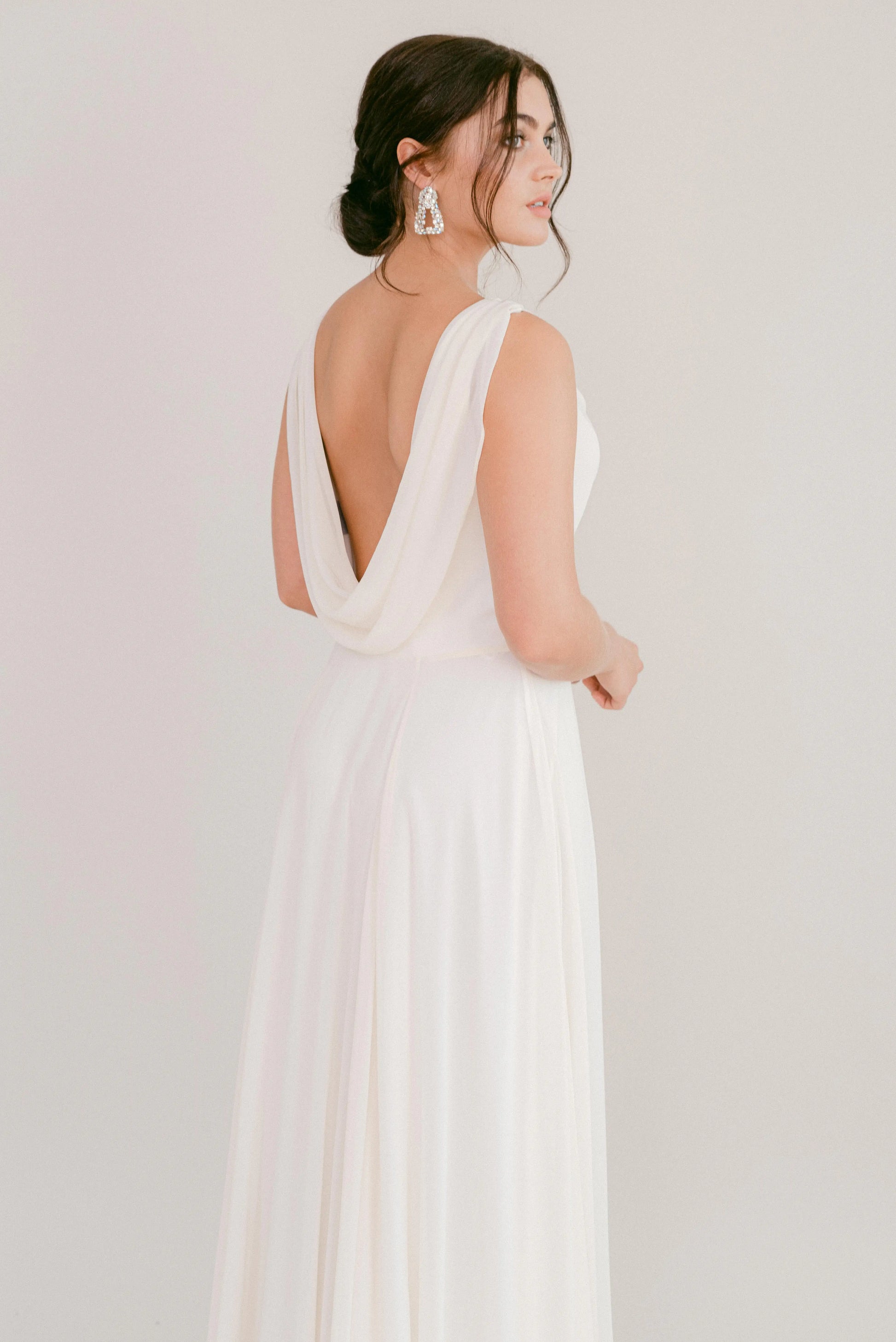 A woman in the TH&TH Athena Dress in Ivory Chiffon stands sideways, gazing over her shoulder. She wears large sparkling earrings and has dark hair styled in an updo, set against a plain, light background.