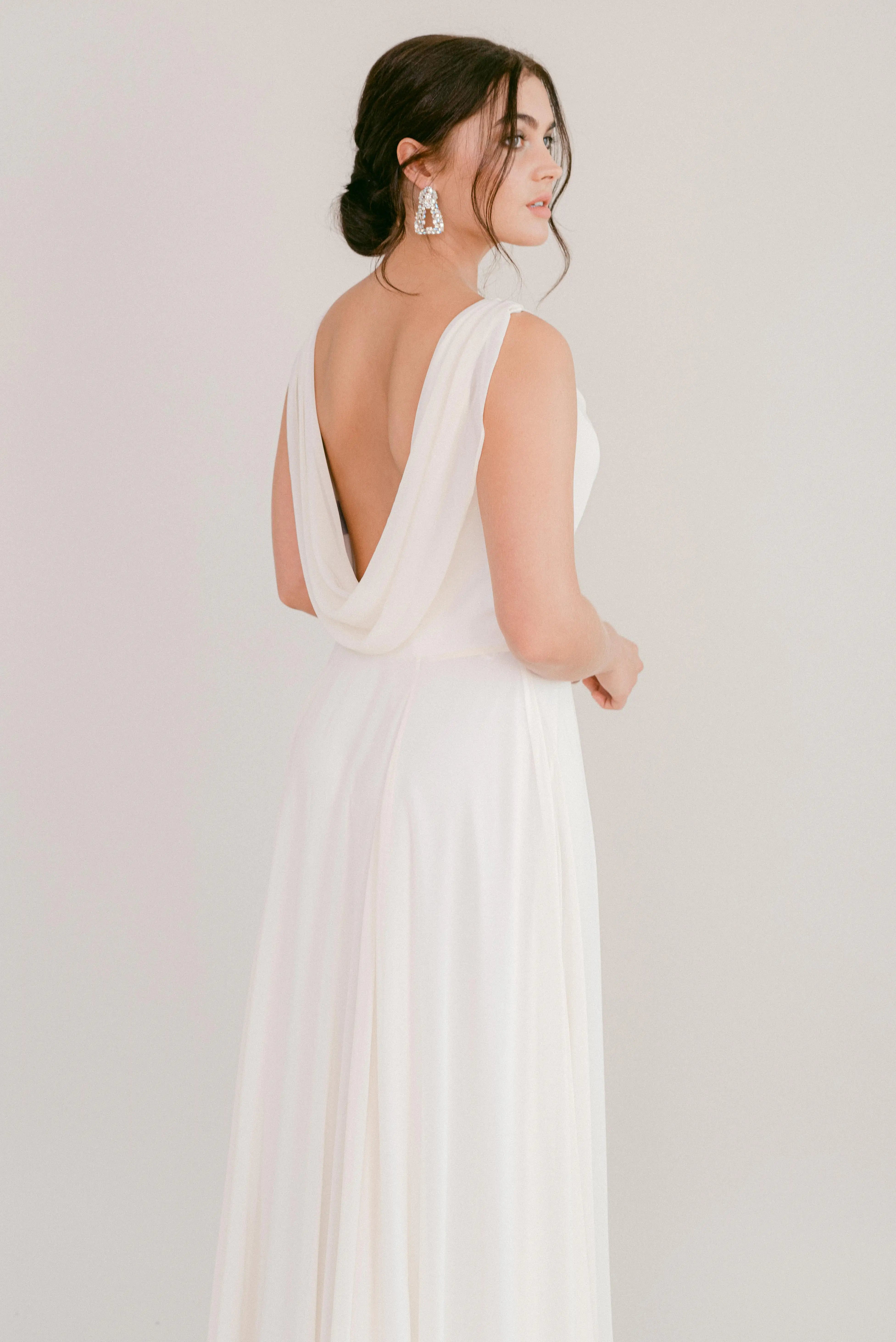 A woman in the TH&TH Athena Dress in Ivory Chiffon stands sideways, gazing over her shoulder. She wears large sparkling earrings and has dark hair styled in an updo, set against a plain, light background.