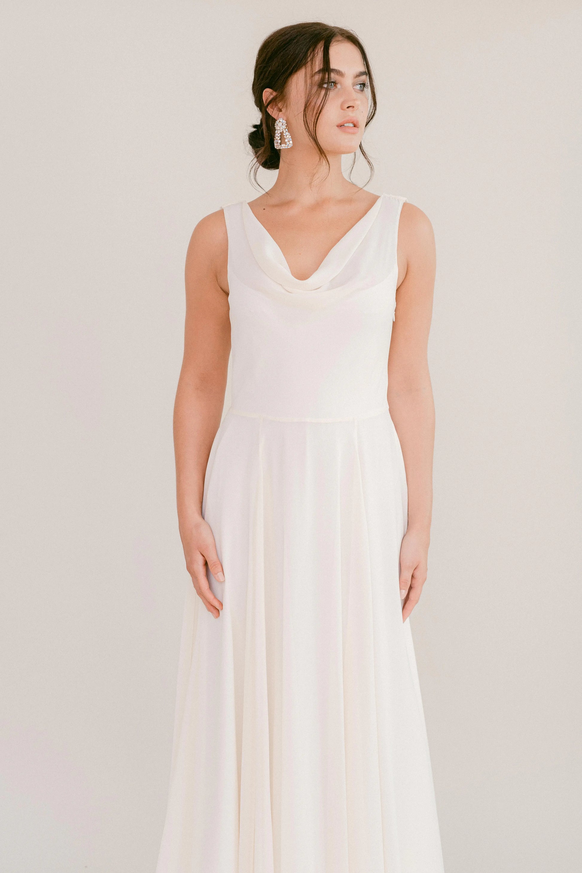 A woman in the TH&TH Athena Dress In Ivory Chiffon, featuring a floor-length silhouette and draped cowl neck, stands against a light background. Her dark hair is styled up, and she accessorizes with statement earrings.