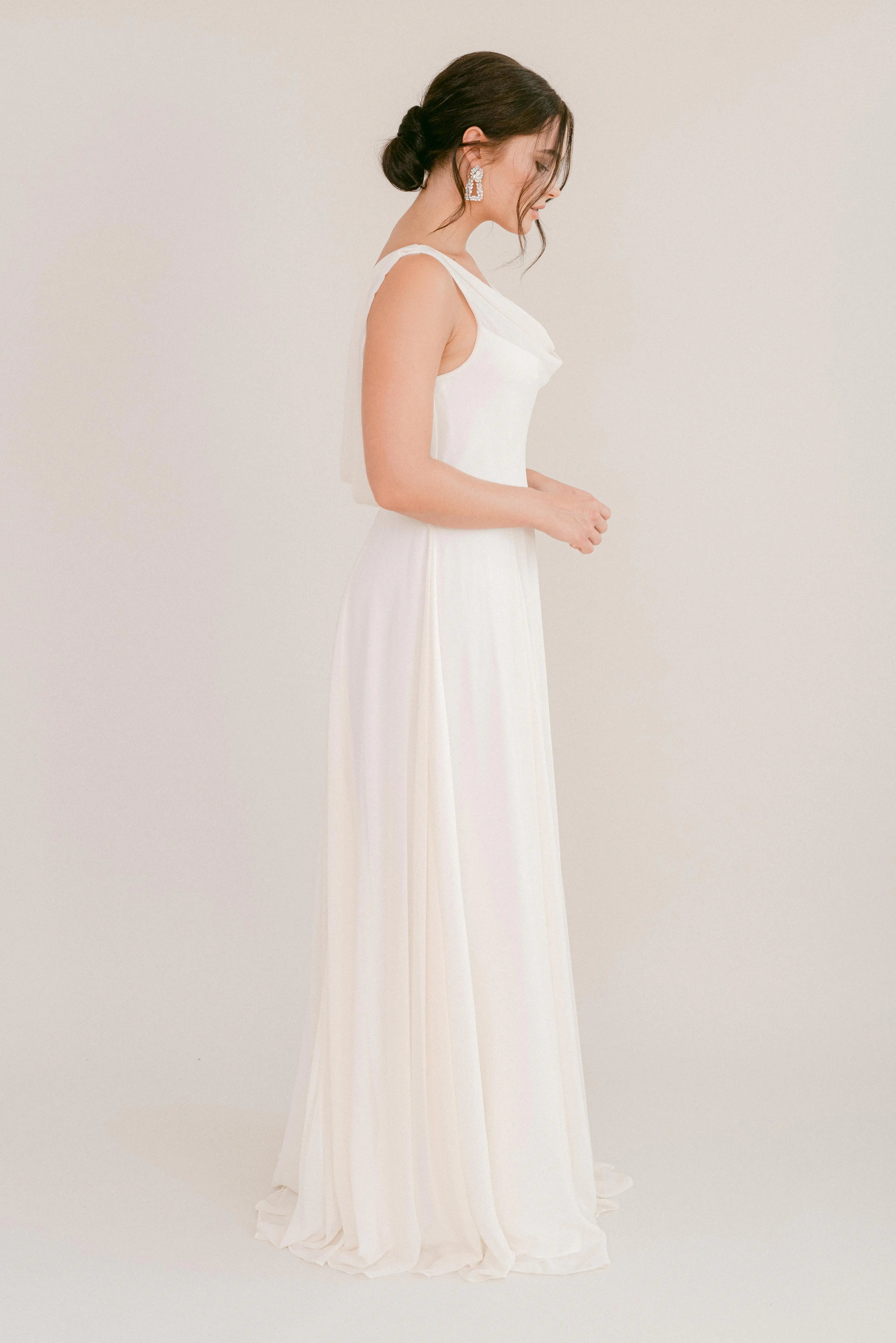 A woman stands in profile against a plain background wearing the TH&TH Athena Dress in Ivory Chiffon, a floor-length, sleeveless bridesmaid gown. Her hair is styled in a low bun, complemented by large statement earrings.