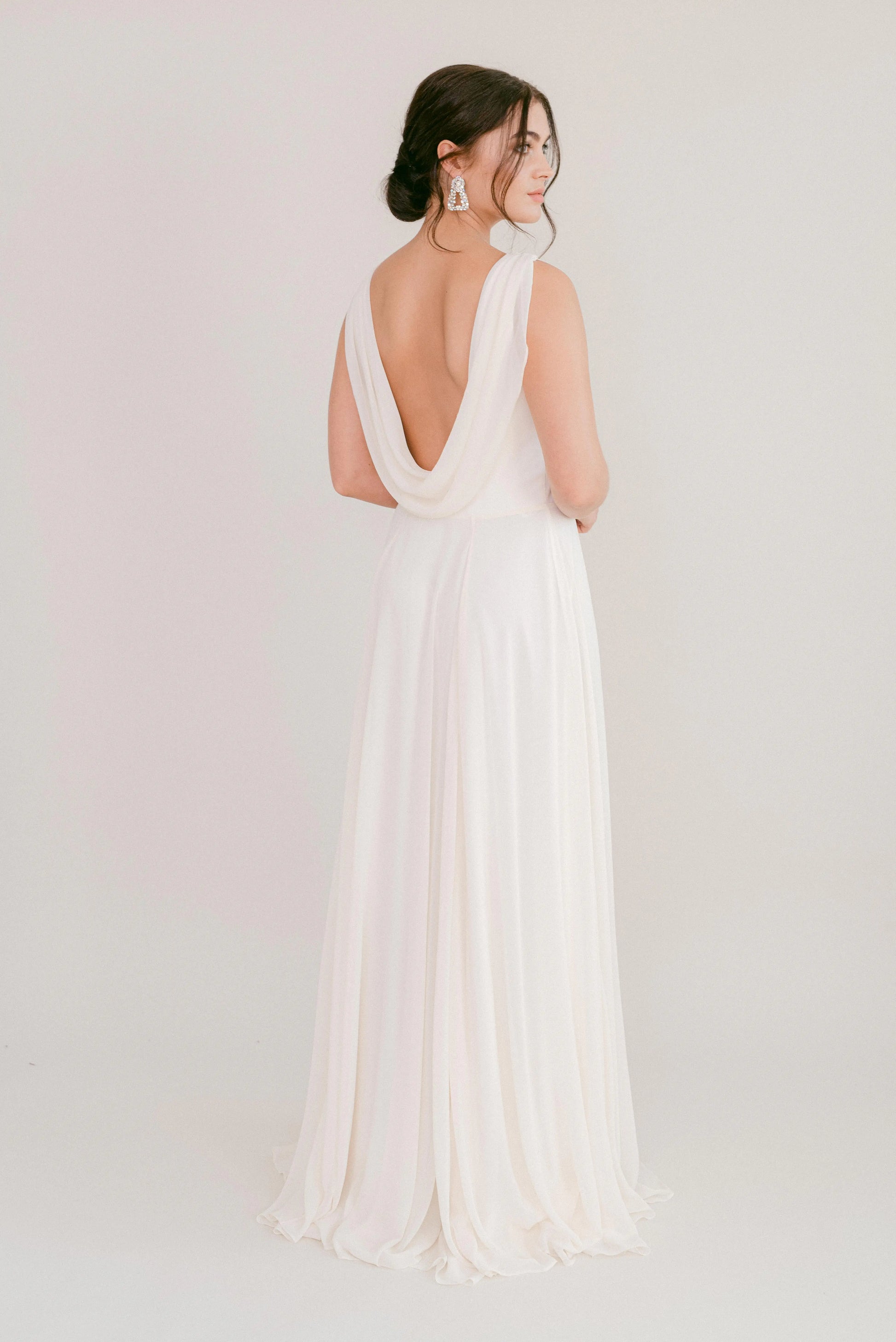 A woman wearing the TH&TH Athena Dress in Ivory Chiffon, featuring a draped low-cut back, stands facing away with her dark hair in a loose updo and statement earrings, set against a plain light background.