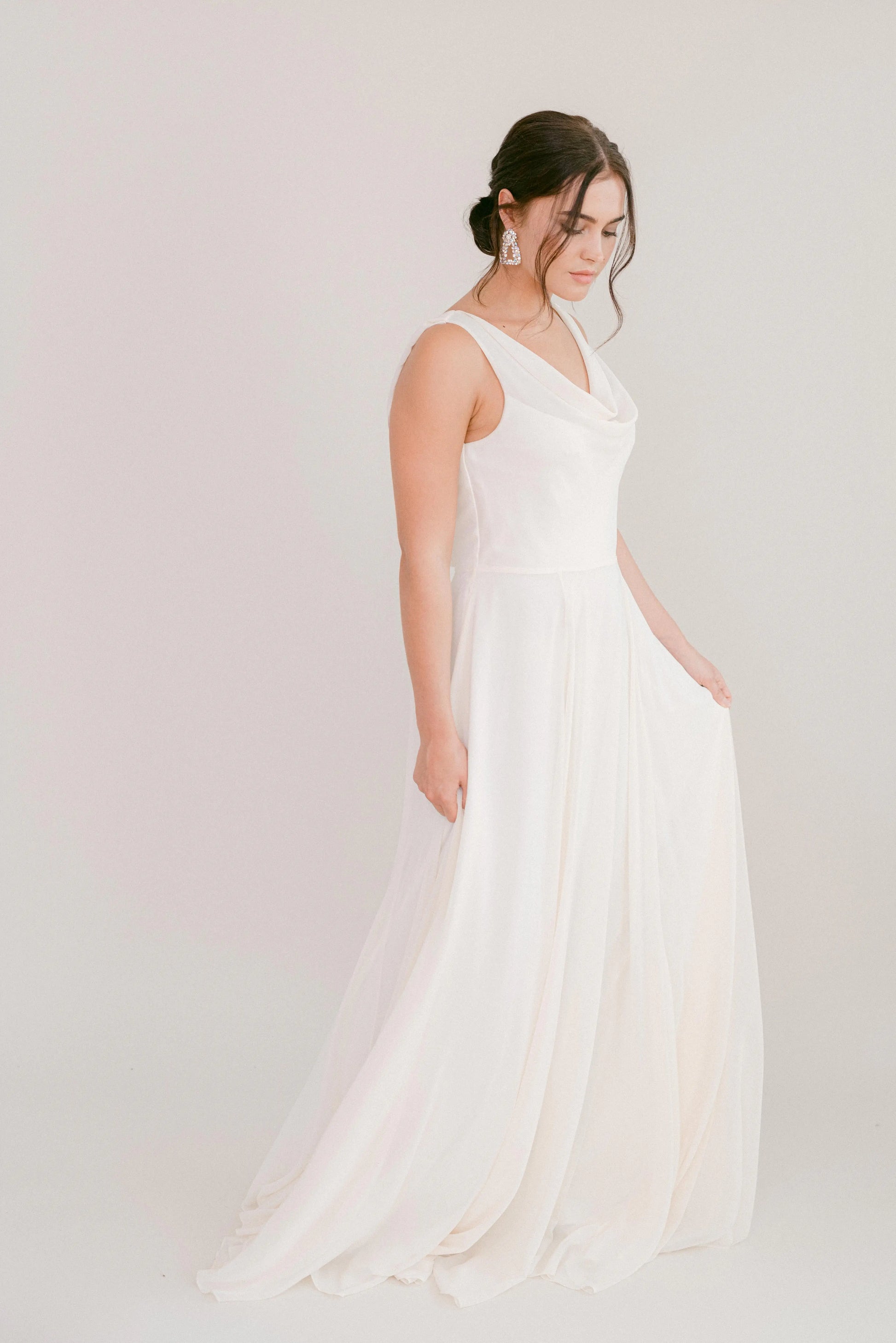 A woman with dark hair in an updo and drop earrings stands against a plain light background, gently holding her TH&TH Athena Dress in Ivory Chiffon as she looks down.