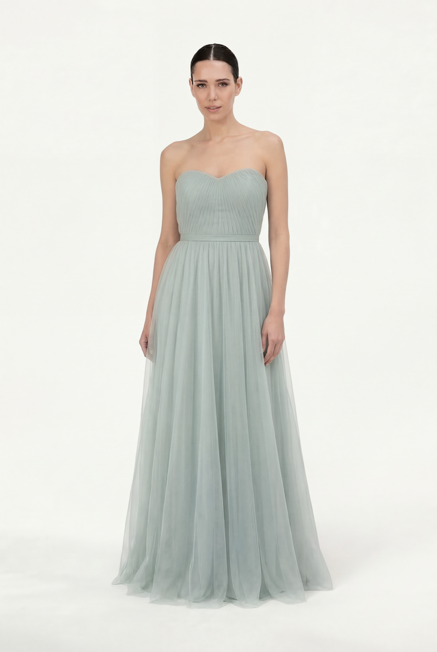 A woman faces forward in the TH&TH Bardot Bridesmaid Dress in Dusty Eucalyptus Tulle, featuring a strapless fitted bodice and flowing pleated skirt against a plain white background.