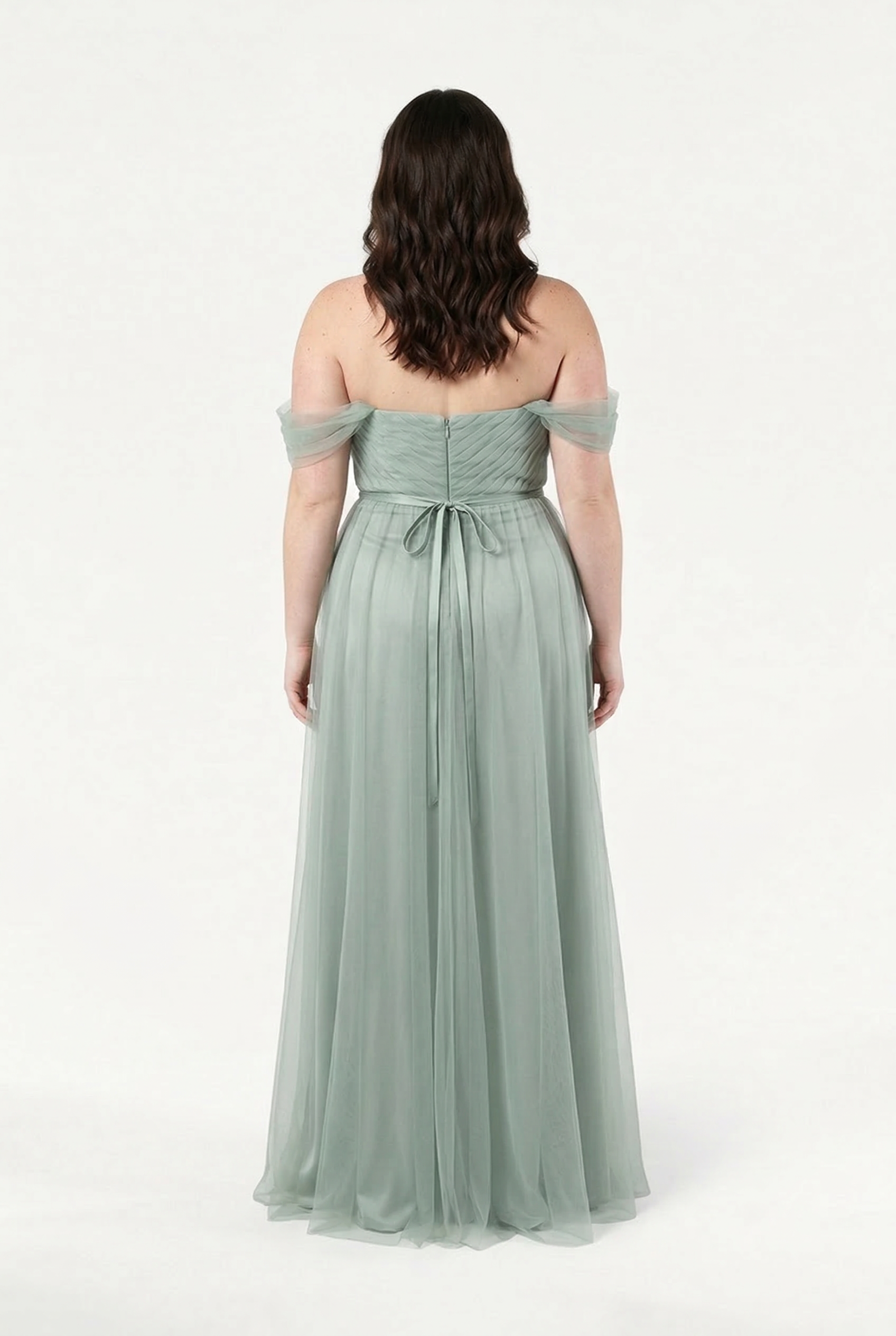 A woman faces away in the TH&TH Bardot Bridesmaid Dress in Dusty Eucalyptus Tulle, featuring an off-shoulder bardot style, tied waist, and flowing skirt. Her wavy brown hair cascades down against a plain white background.