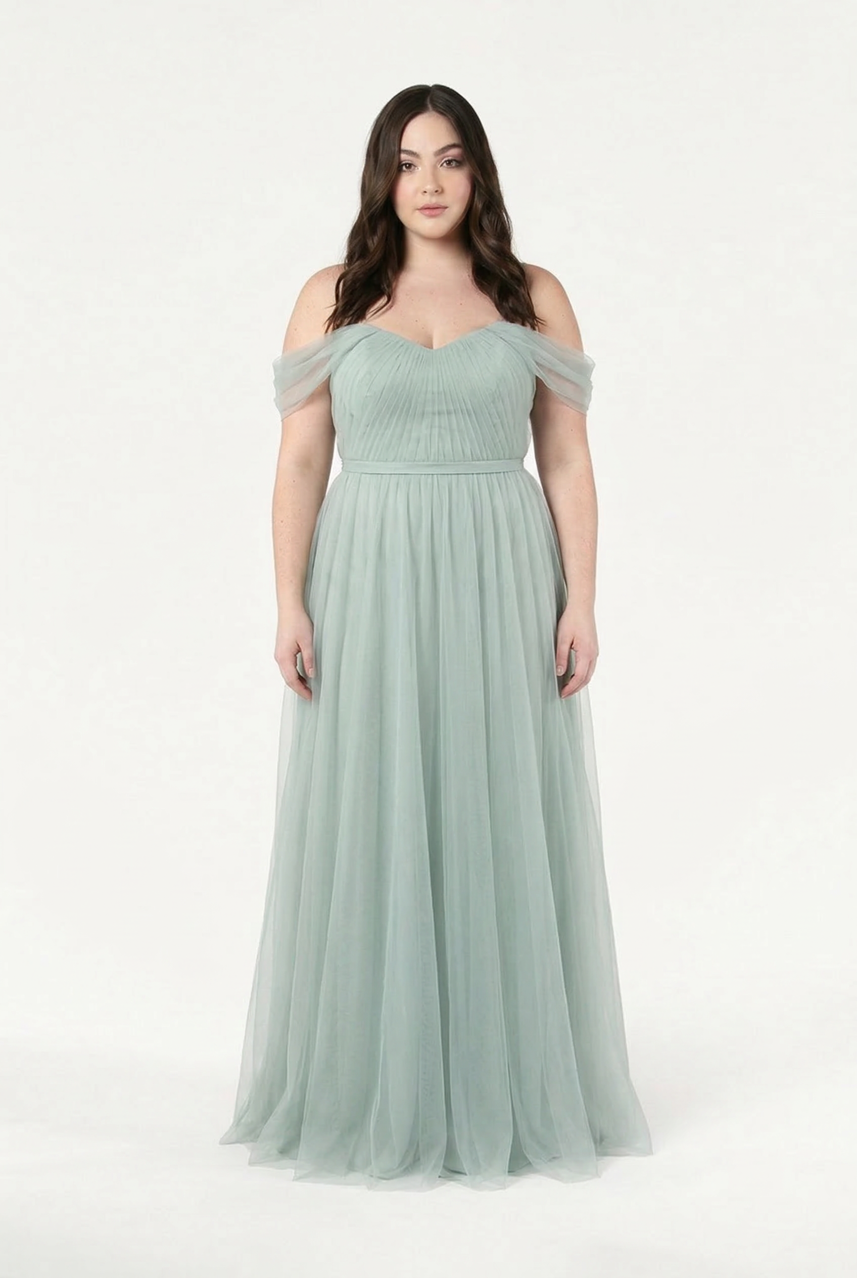 A woman with long, wavy brown hair wears the TH&TH Bardot Bridesmaid Dress in Dusty Eucalyptus Tulle, featuring an off-shoulder design and fitted bodice, standing against a plain white background.