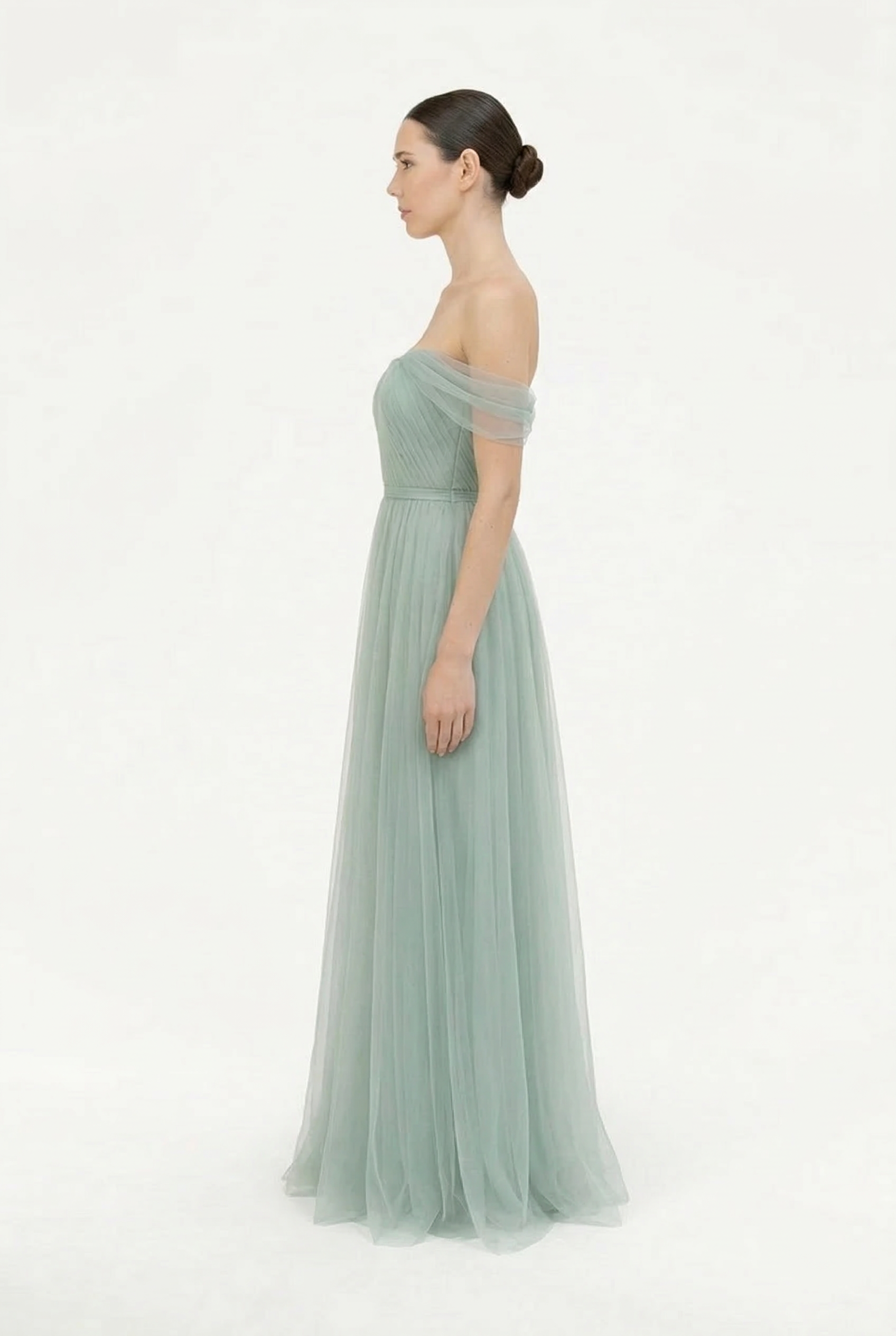 A woman in profile wears the TH&TH Bardot Bridesmaid Dress in Dusty Eucalyptus tulle, featuring an off-the-shoulder neckline, fitted bodice, and flowing skirt. Her hair is styled in a neat bun against a plain white background.