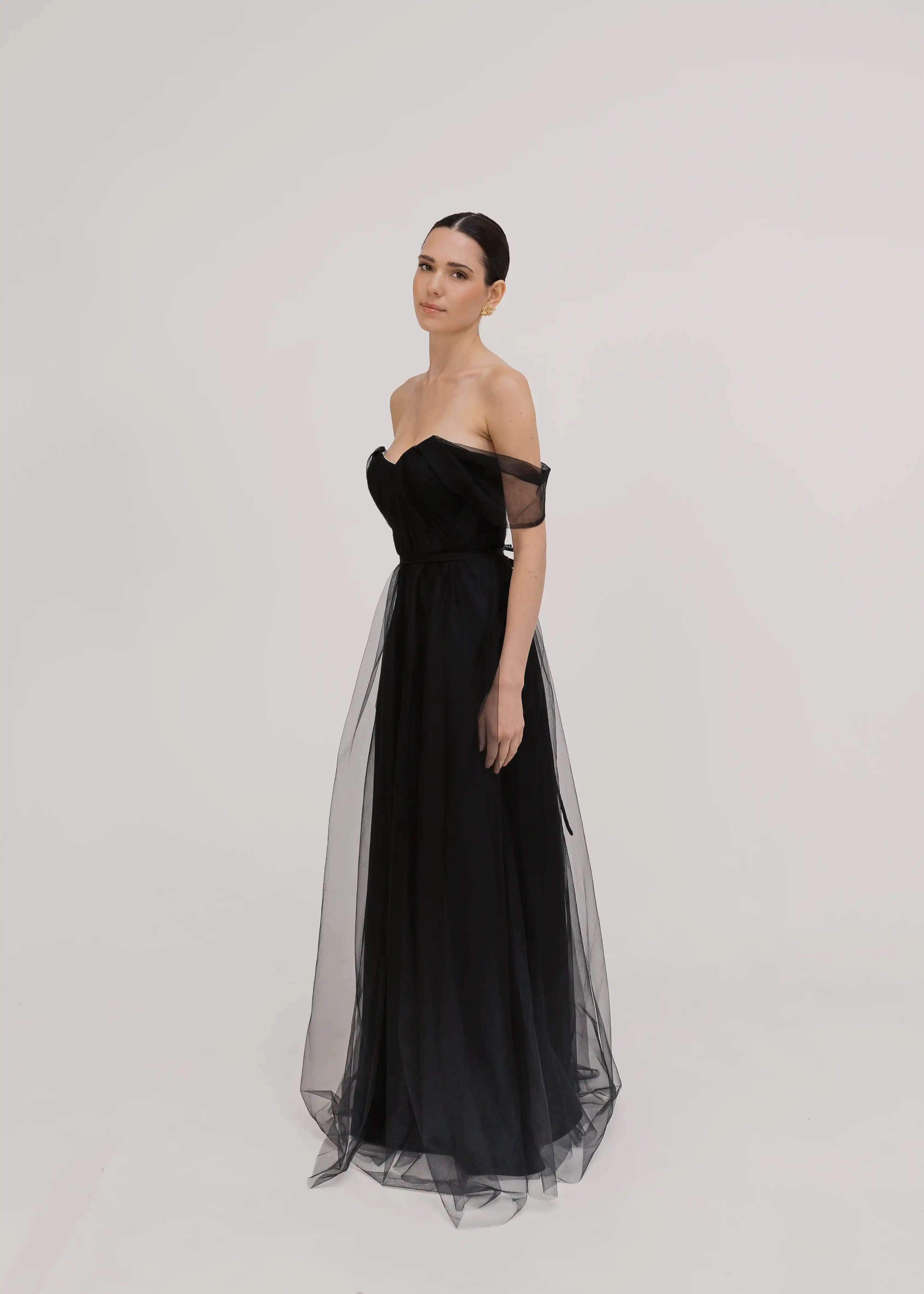 Model wears the TH&TH Bardot Bridesmaid Dress in Black Tulle—a floor-length, off-shoulder gown with sheer overlay—standing against a plain white background, looking slightly to the side with a neutral expression.