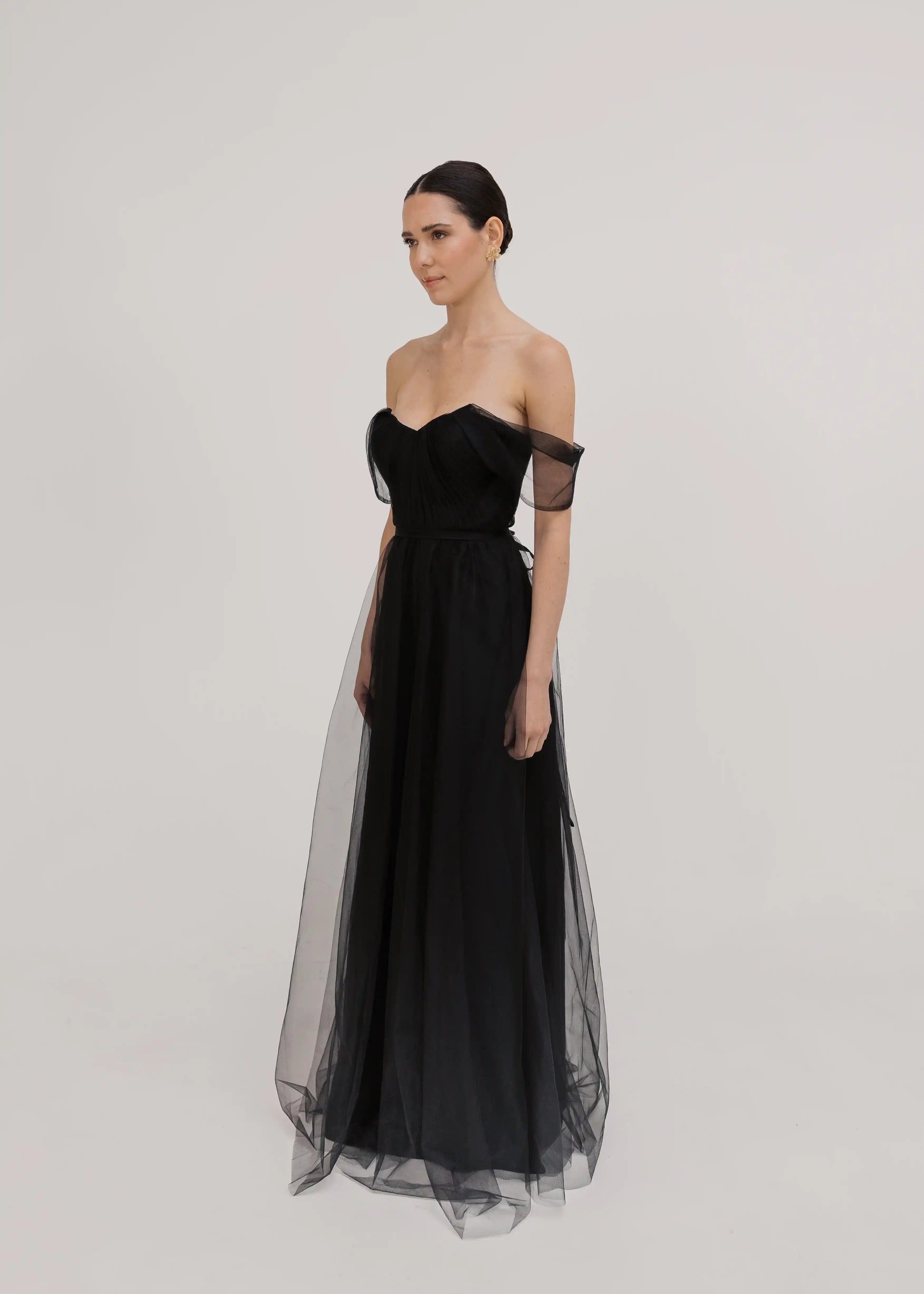A woman poses in a studio, gazing sideways while wearing the TH&TH Bardot Bridesmaid Dress in Black Tulle—an off-shoulder, floor-length gown with a sheer overlay—her dark hair pulled back against a plain light background.