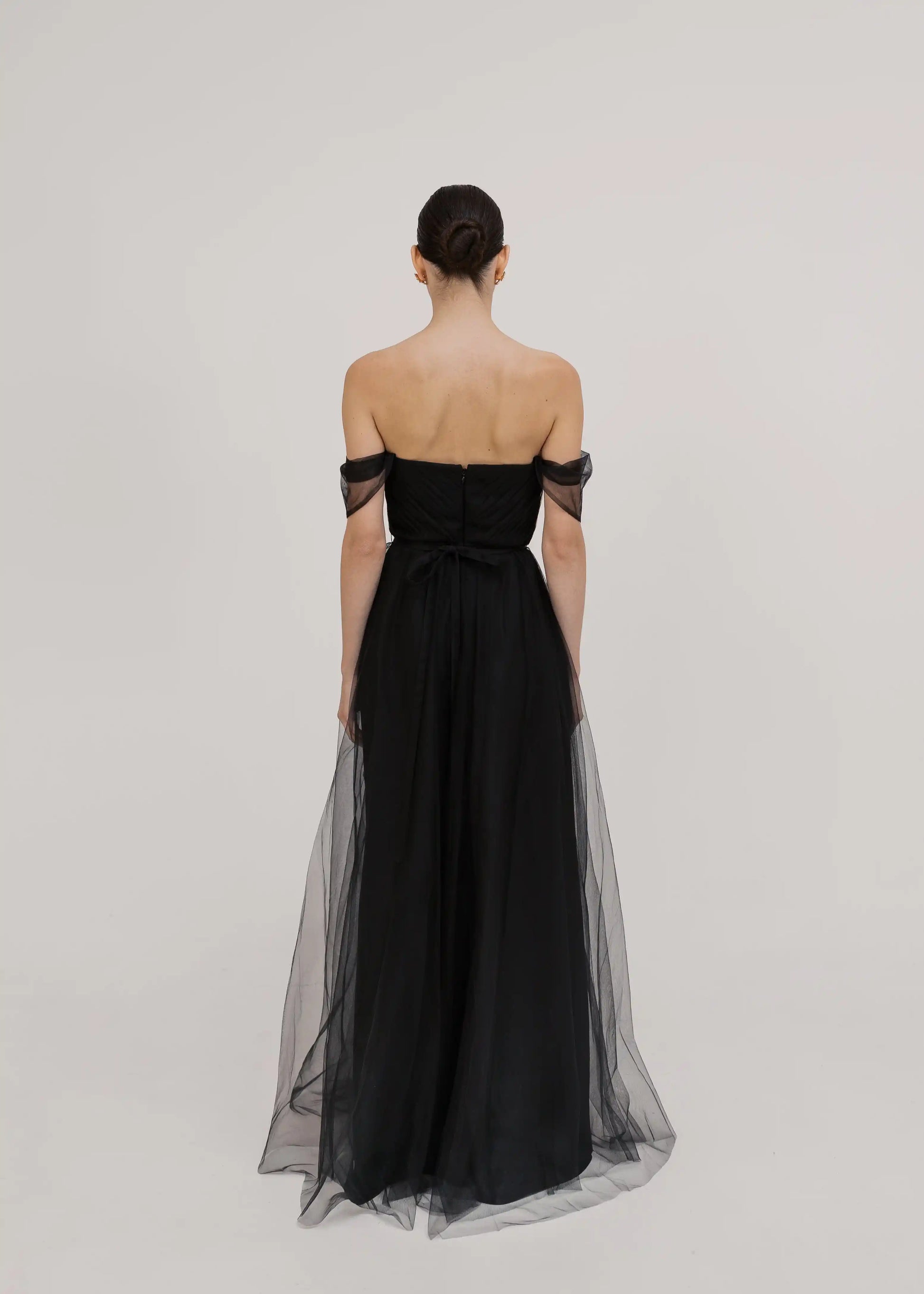 A woman stands with her back to the camera in the TH&TH Bardot Bridesmaid Dress in Black Tulle, featuring a long, strapless design with off-the-shoulder sleeves. Her hair is styled in a neat bun against a plain light background.