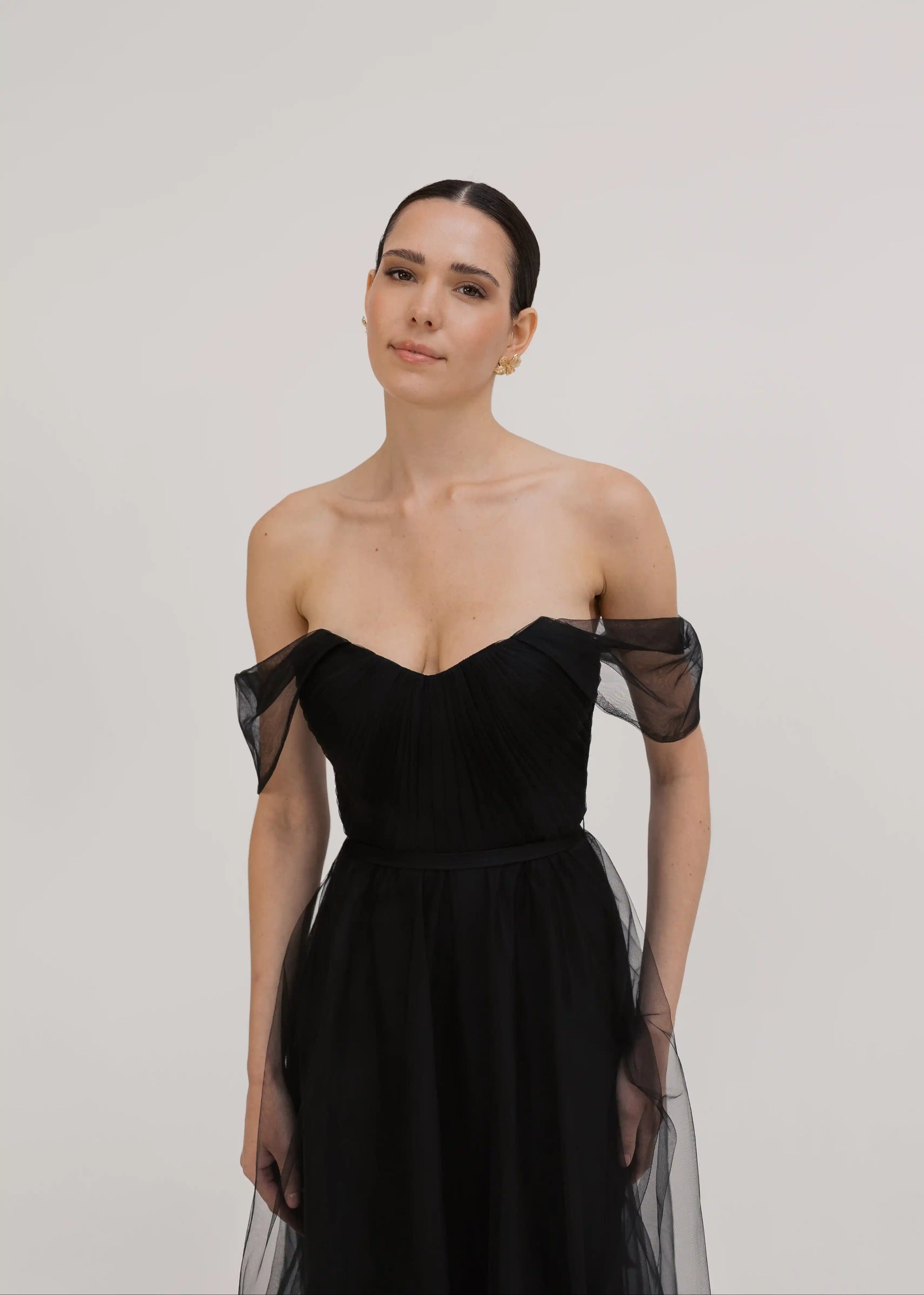 A woman with dark hair pulled back wears the TH&TH Bardot Bridesmaid Dress in Black Tulle, standing against a light background and looking calmly at the camera.