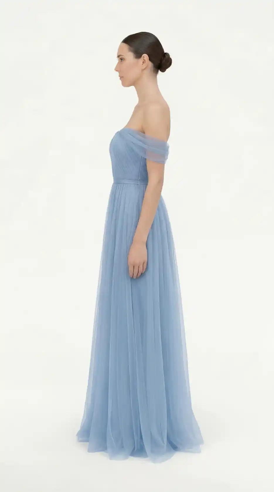 A woman stands in profile wearing the TH&TH Bardot Bridesmaid Dress in Dusty Blue Tulle, featuring a fitted bodice and flowing skirt. Her hair is styled in a neat bun against a plain white background.