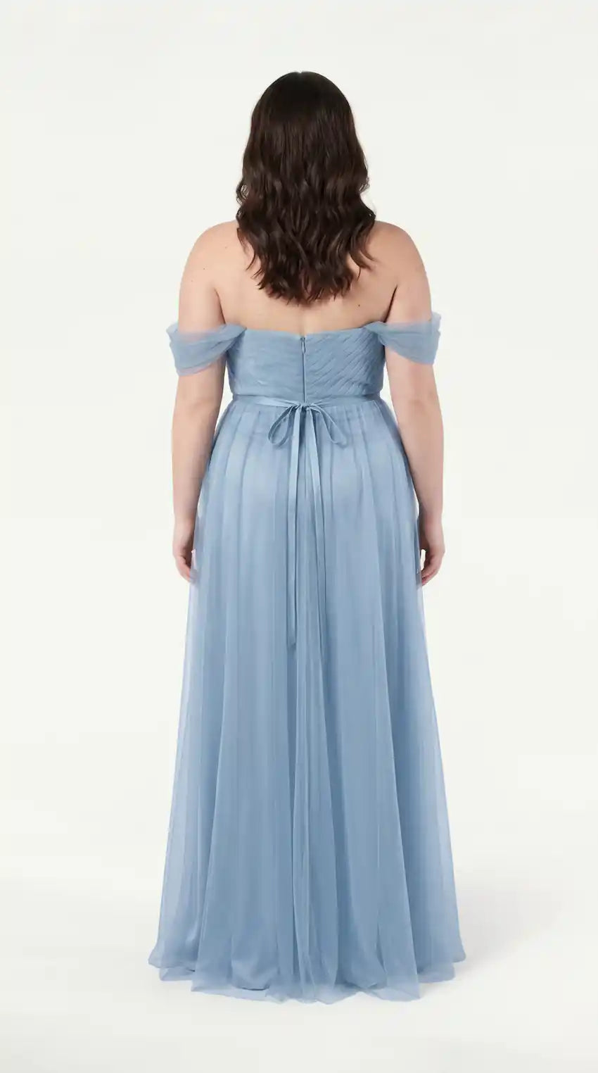 A woman with wavy brown hair faces away, wearing the TH&TH Bardot Bridesmaid Dress in Dusty Blue Tulle—an elegant off-shoulder dress with a fitted bodice and flowing skirt, shown against a plain white background.