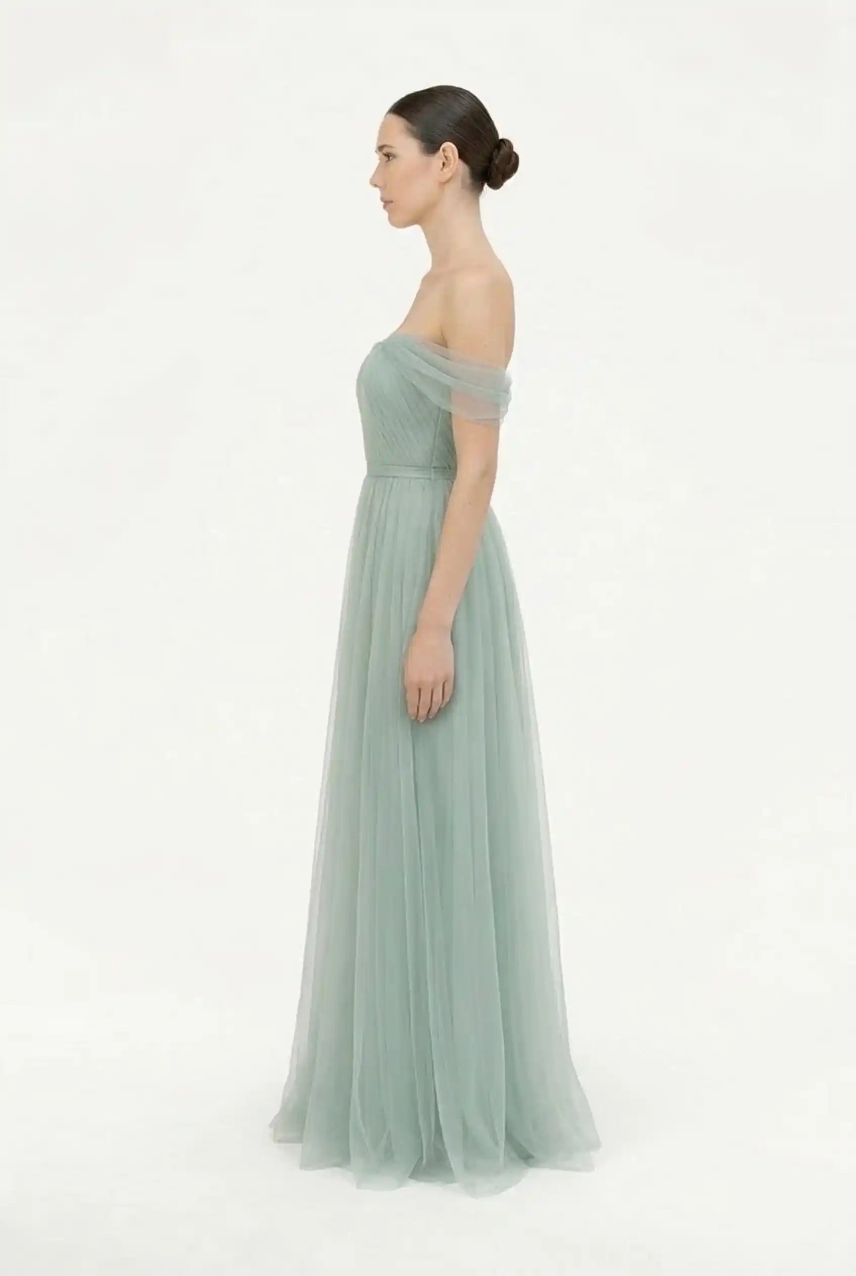 A woman in profile wears the TH&TH Bardot Bridesmaid Dress in Dusty Eucalyptus tulle, featuring an off-the-shoulder neckline, fitted bodice, and flowing skirt. Her hair is styled in a neat bun against a plain white background.