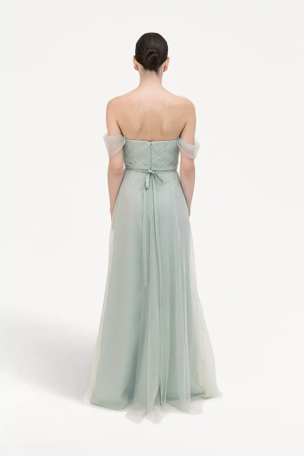 A woman with neatly pulled-back dark hair faces away, wearing the TH&TH Bardot Bridesmaid Dress in Dusty Eucalyptus Tulle, featuring sheer, flowing fabric and a tied waist sash, set against a plain white background.